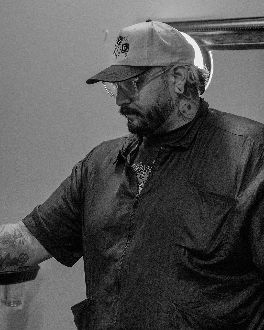 Black and white photo of a man with tattoos, beard, glasses, wearing a baseball cap, black jacket, and T-shirt, looking down.