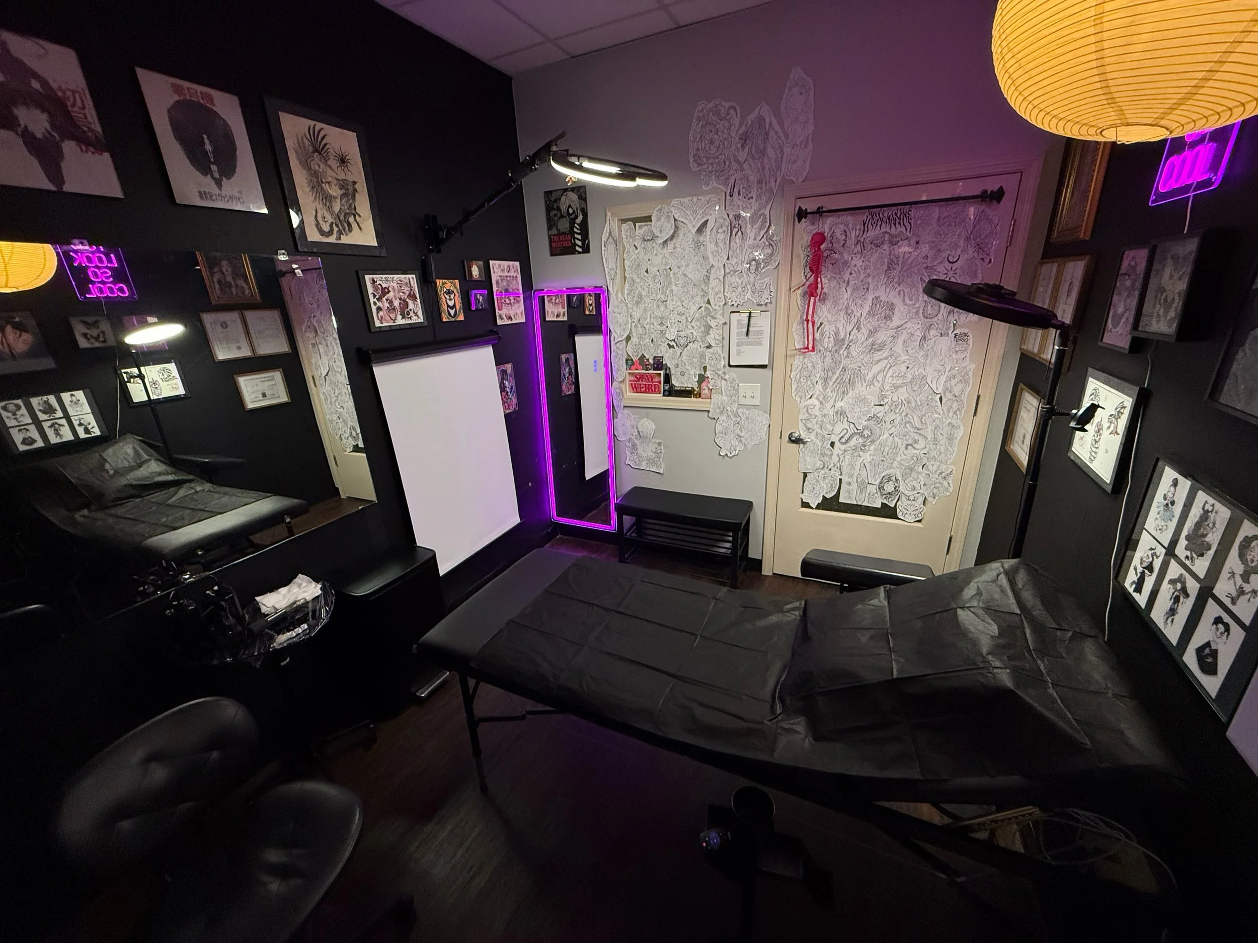 Tattoo studio or art studio room with black walls decorated with framed artwork, a tattoo chair covered in black paper, and various art pieces on the walls, illuminated by neon and soft lighting.