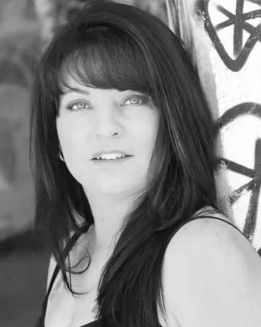 Black and white photo of a woman with long dark hair, light eyes, and a confident expression, standing outdoors near a graffiti-covered wall.