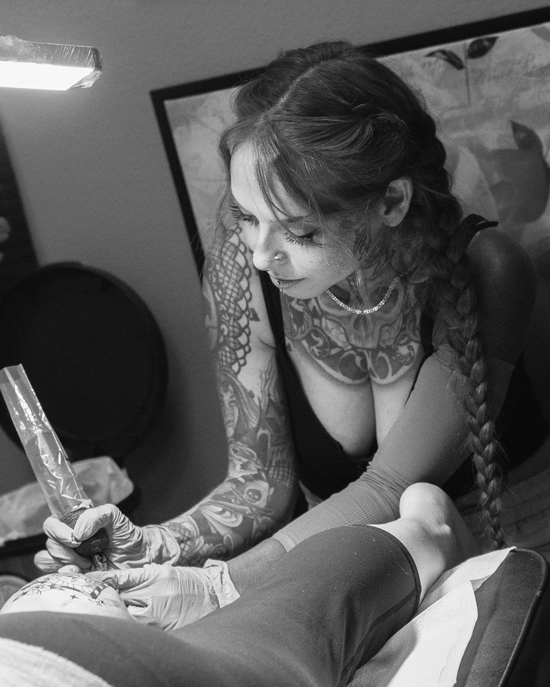 A woman with tattoos and long braided hair, wearing gloves, is tattooing a person's arm in a studio.
