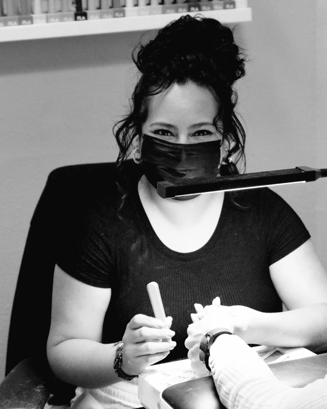 A woman wearing a black face mask, sitting at a table, signing something with a pen, with people in the background.