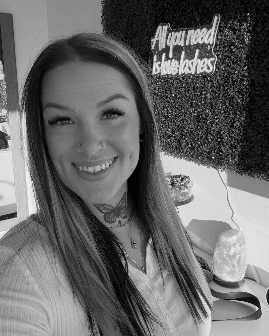 A smiling woman with long hair and a butterfly tattoo on her neck, standing in front of a textured wall with a neon sign that reads 'All you need is eyelash'.