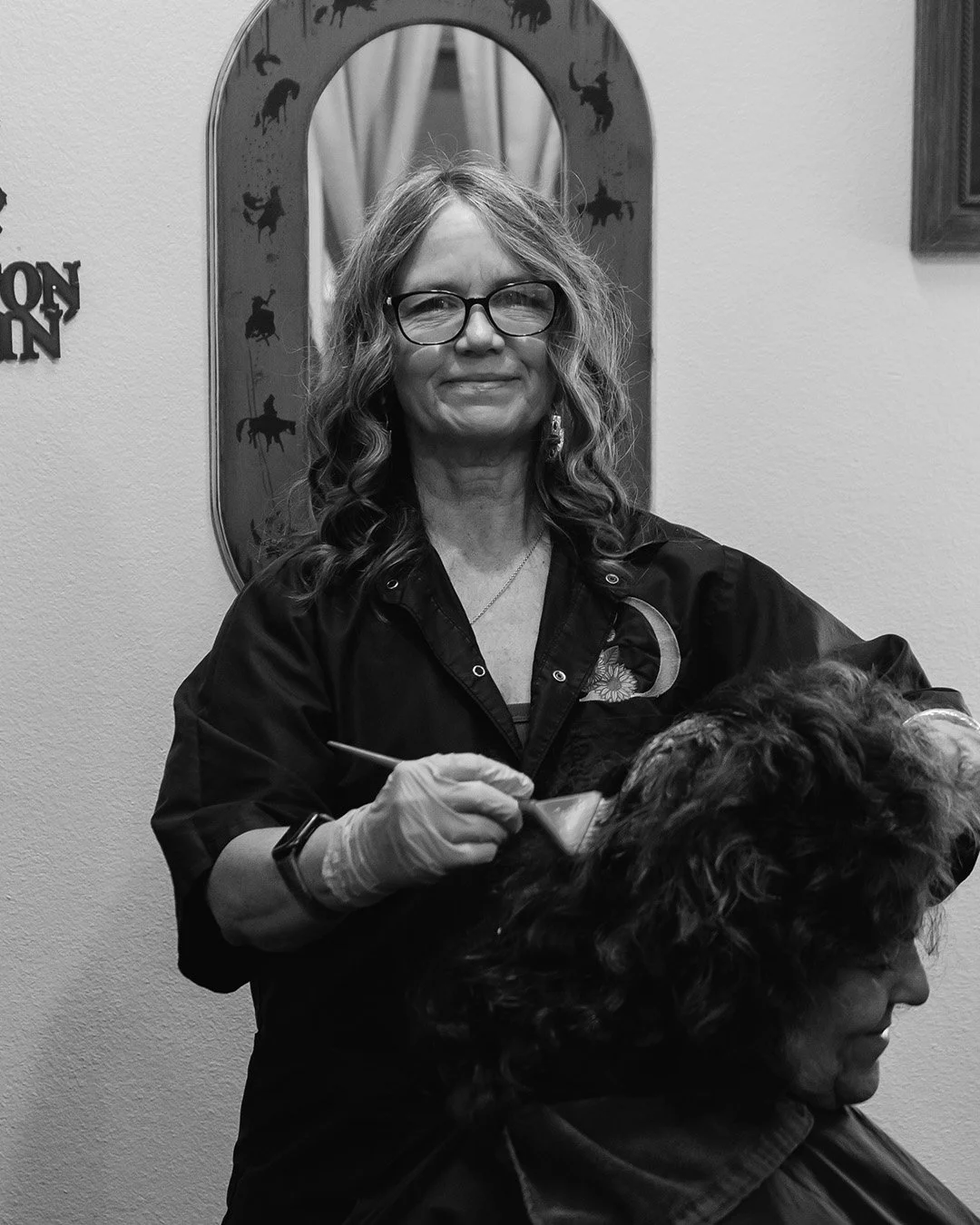 A woman with glasses and curly hair giving a haircut to another woman with curly hair in a salon, with a mirror and wall decorations in the background.
