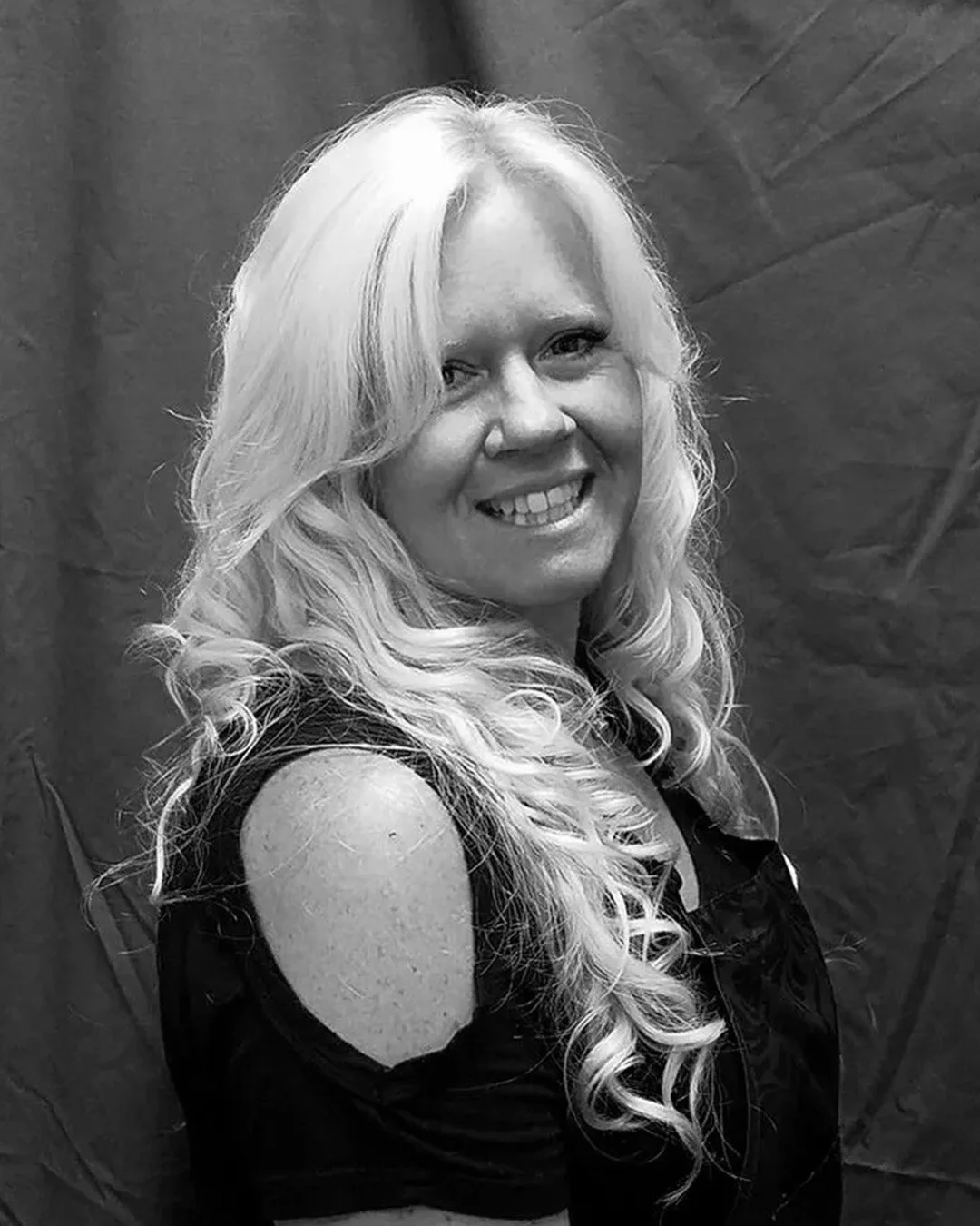 Black and white photo of a woman with long, curly, light-colored hair smiling and looking at the camera, wearing a sleeveless top with a cold shoulder design.