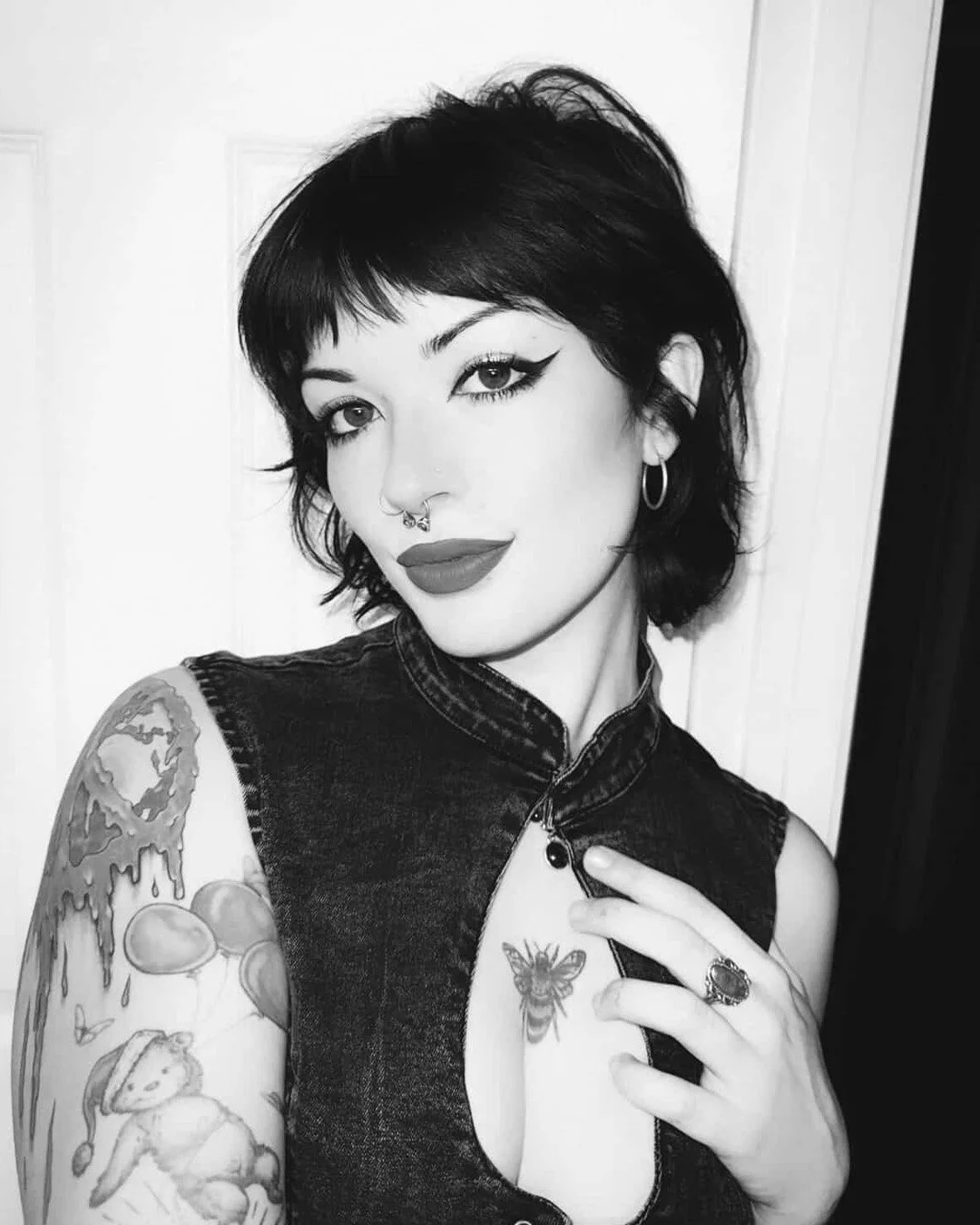 A woman with short dark hair, wearing a sleeveless denim top, has tattoos on her arm and chest, including a butterfly. She has makeup with winged eyeliner, bold lipstick, earrings, and nose piercings, and is taking a selfie in front of a plain background.