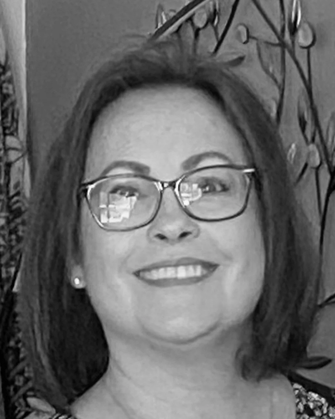 A woman with glasses, shoulder-length hair, and earrings, smiling in front of houseplants and floral decor