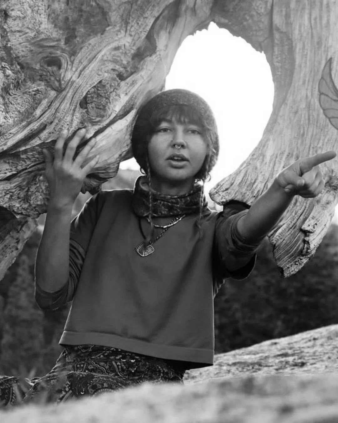A person with short hair, wearing a necklace and a long-sleeved shirt, is holding a large piece of wood or tree branch over their shoulders and pointing with their right hand. The photo is in black and white.