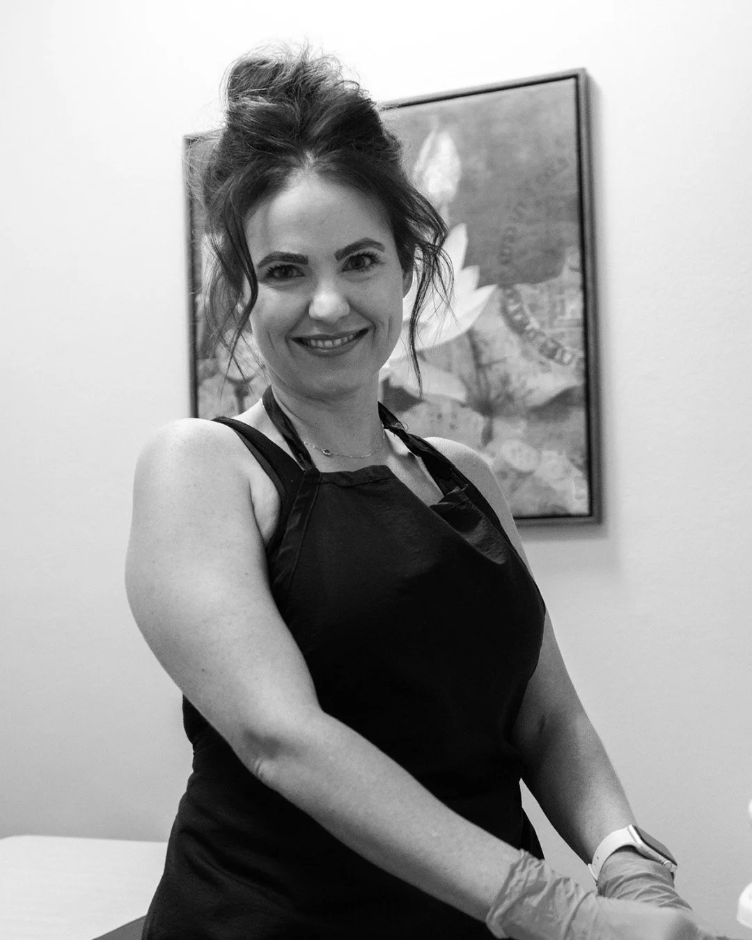 A smiling woman with dark, wavy hair in an updo, wearing a sleeveless black top and gloves, standing in front of a framed artwork on a plain wall.