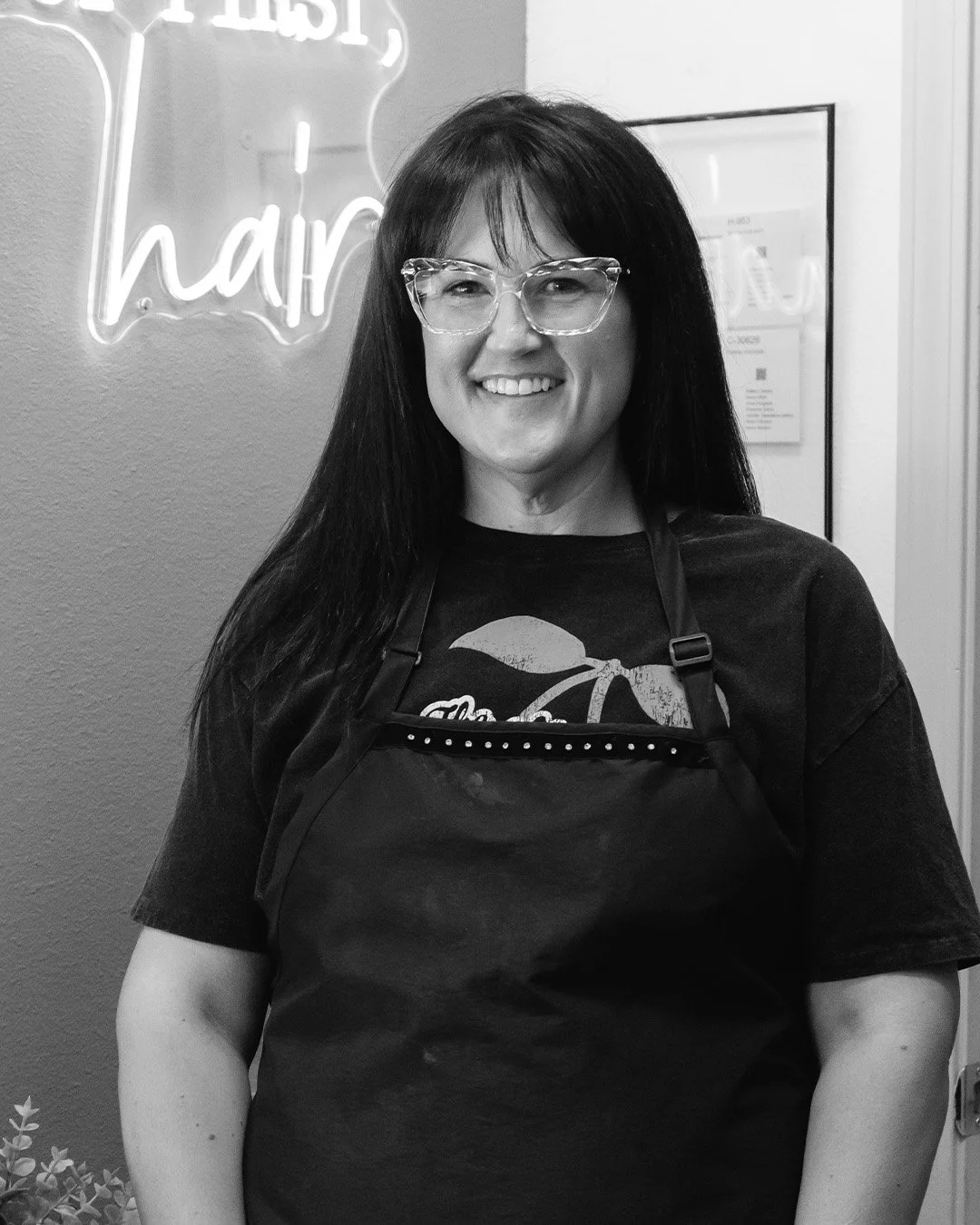A woman with long dark hair, glasses, and a smile, wearing a black apron, standing in front of a wall with a neon sign that says 'hair'.
