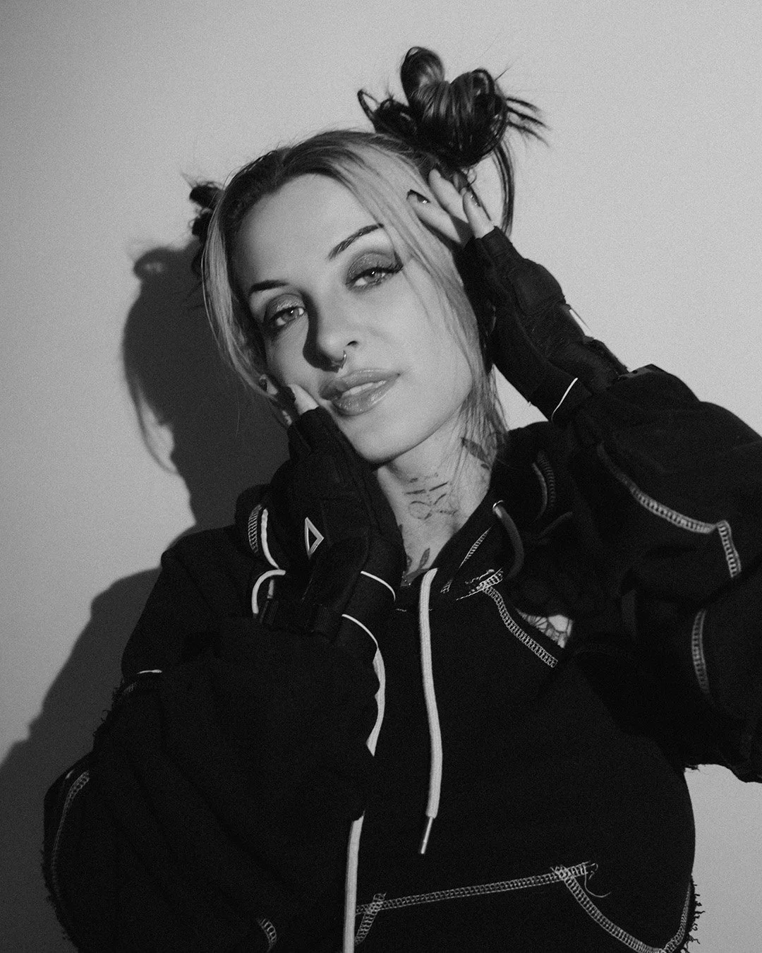 Black and white photo of a woman with light hair styled in buns, wearing a black hoodie with zippered pocket details, holding her face with both hands, and looking at the camera.