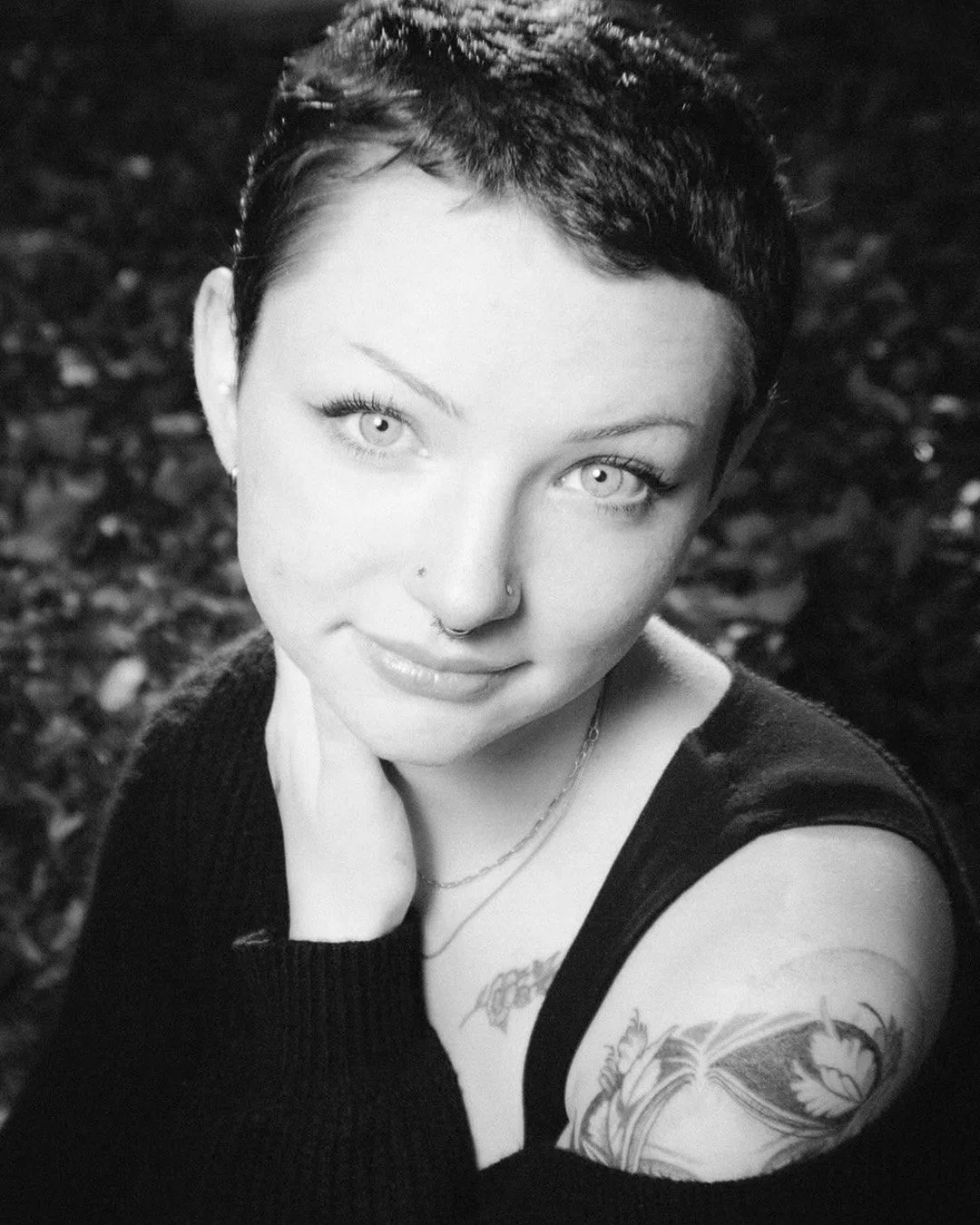Black and white close-up portrait of a woman with short dark hair, tattoos on her shoulder and chest, and piercing eyes, smiling softly and looking at the camera.