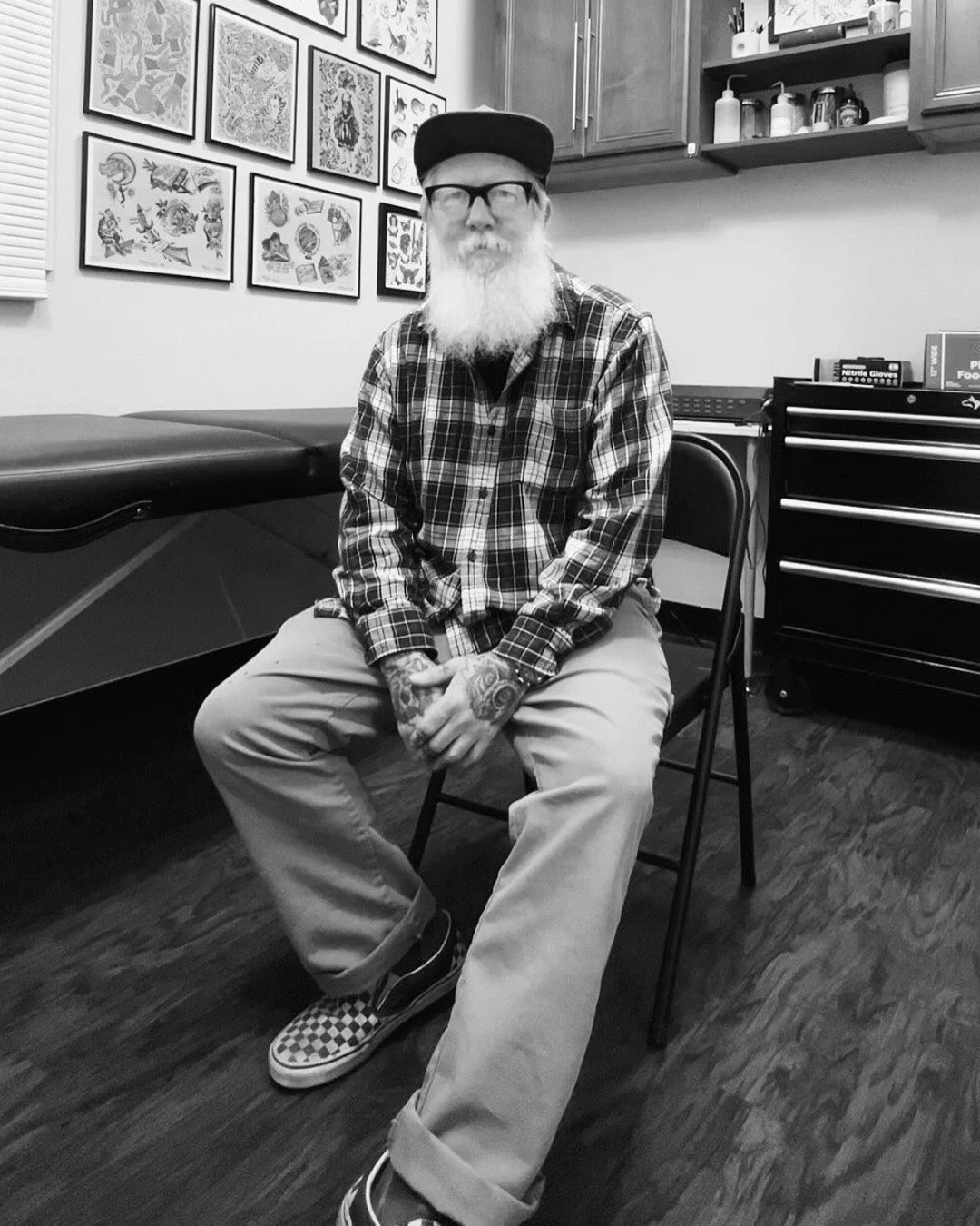 A man with glasses, a cap, a long beard, and tattoos on his arms, sitting on a chair in a tattoo shop. He is wearing a plaid shirt, khaki pants, and checkered slip-on shoes. Behind him are framed artwork on the wall, a tattoo chair, cabinets, and a toolbox.