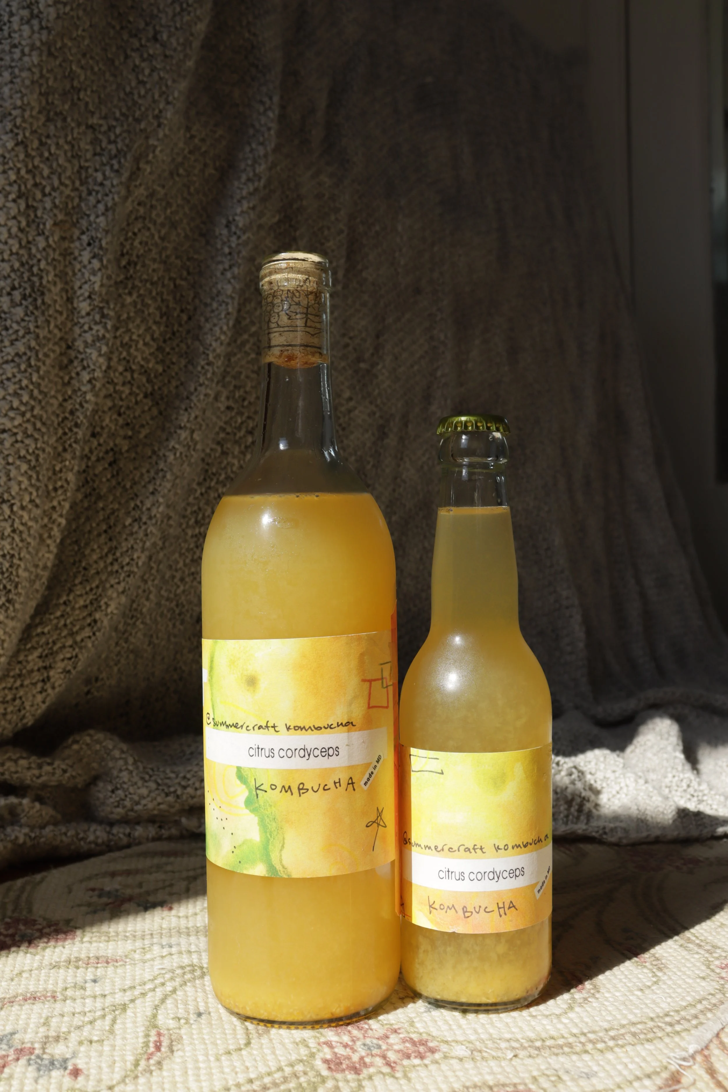 Two bottles of homemade kombucha with yellow-colored liquid, labels indicating flavor as citrus cordyceps, placed on a textured cloth surface with a fuzzy background.