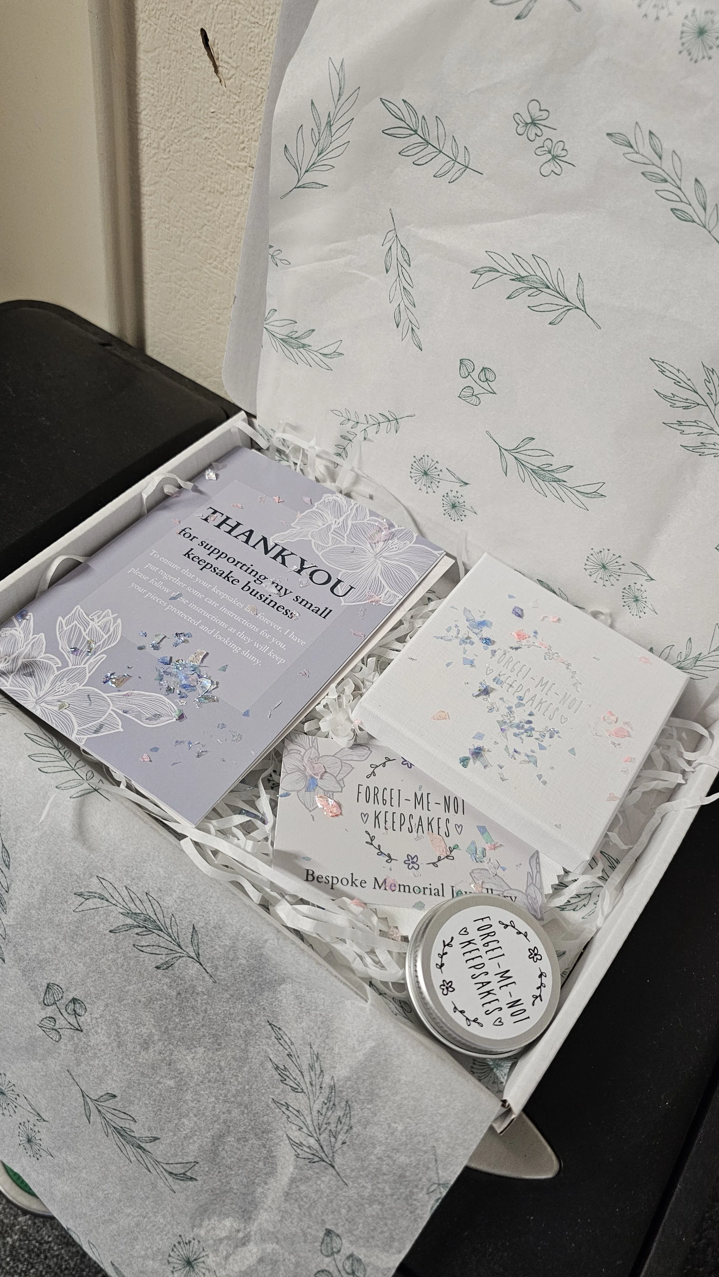 A white box with botanical print paper lining contains small cards, a tin container, and a small white box, all decorated with floral and leaf designs, and some with glitter accents.