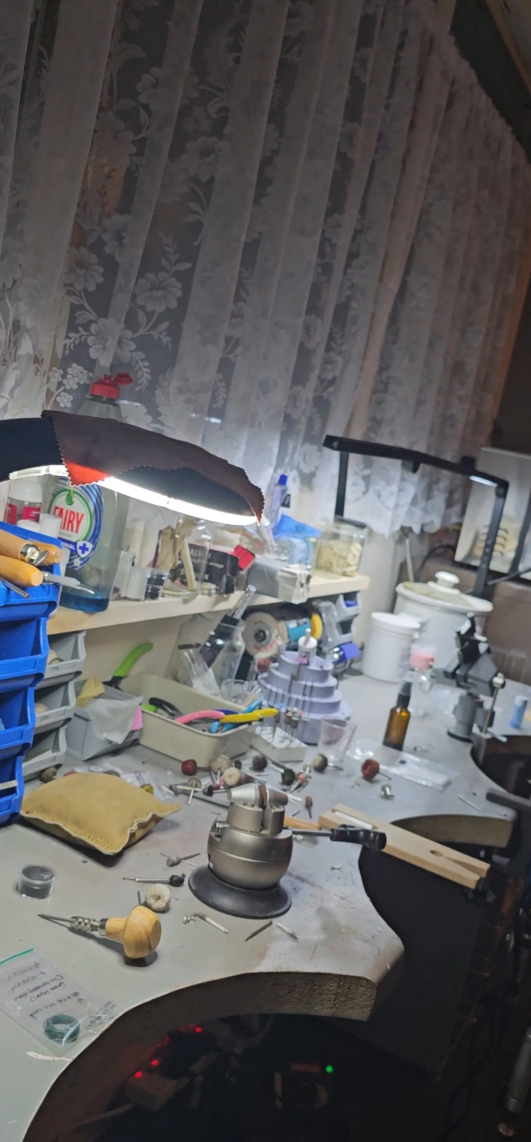 A cluttered workbench with jewelry-making tools, supplies, and equipment, illuminated by a bright lamp, with lace curtains in the background.