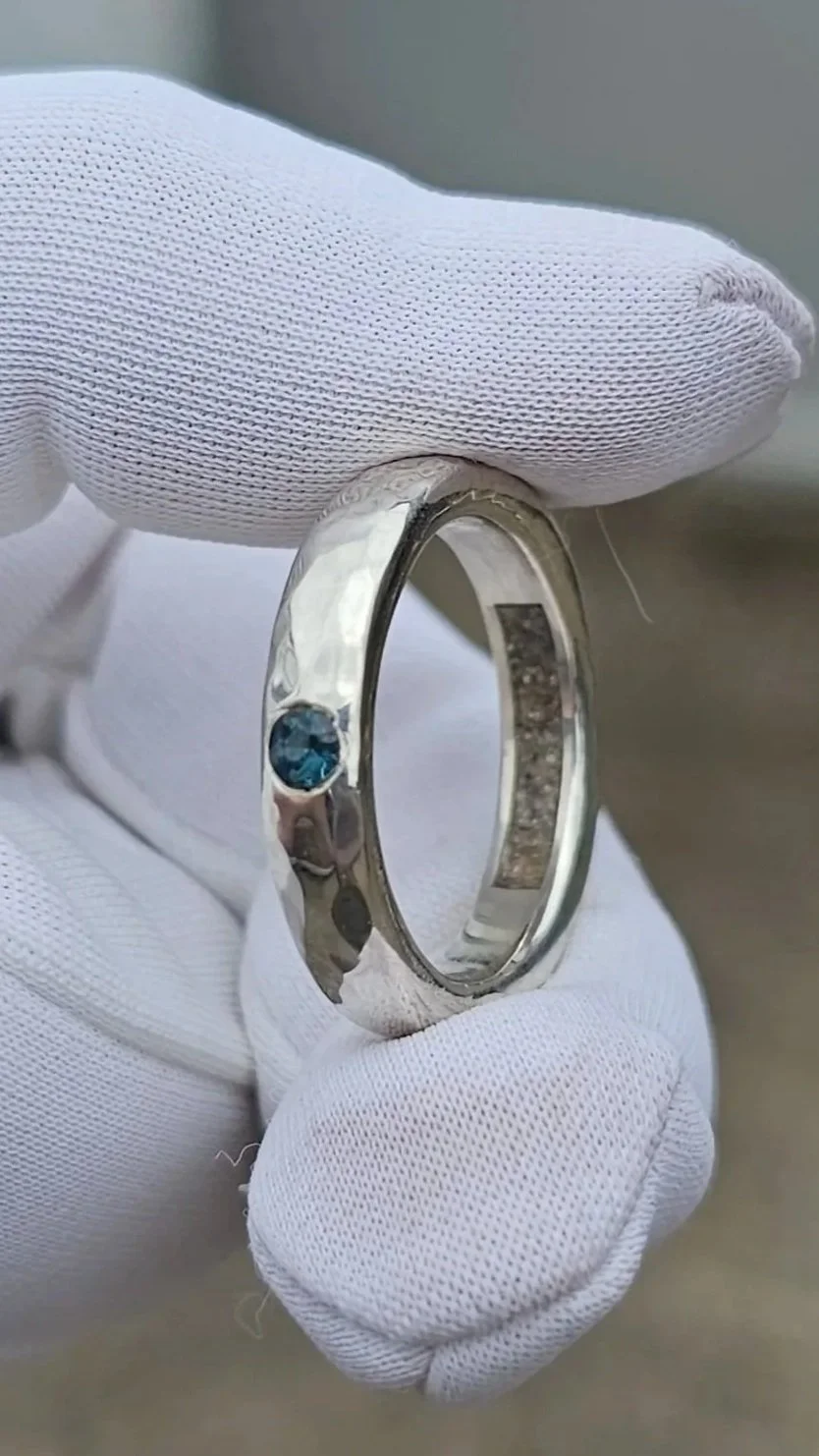 A person wearing white gloves holding a silver ring with a small blue gemstone in a bezel setting.