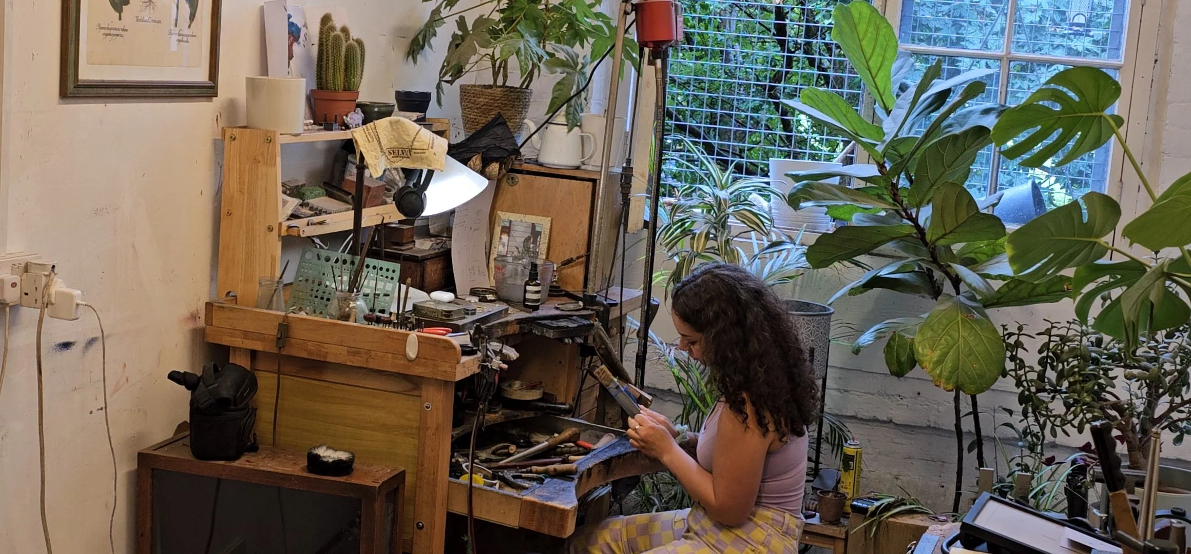 A woman with curly hair wearing a pink tank top and yellow checkered pants sitting on a small stool in an indoor workshop or studio, working on a tool or piece of jewelry, surrounded by various tools, plants, and workshop supplies.