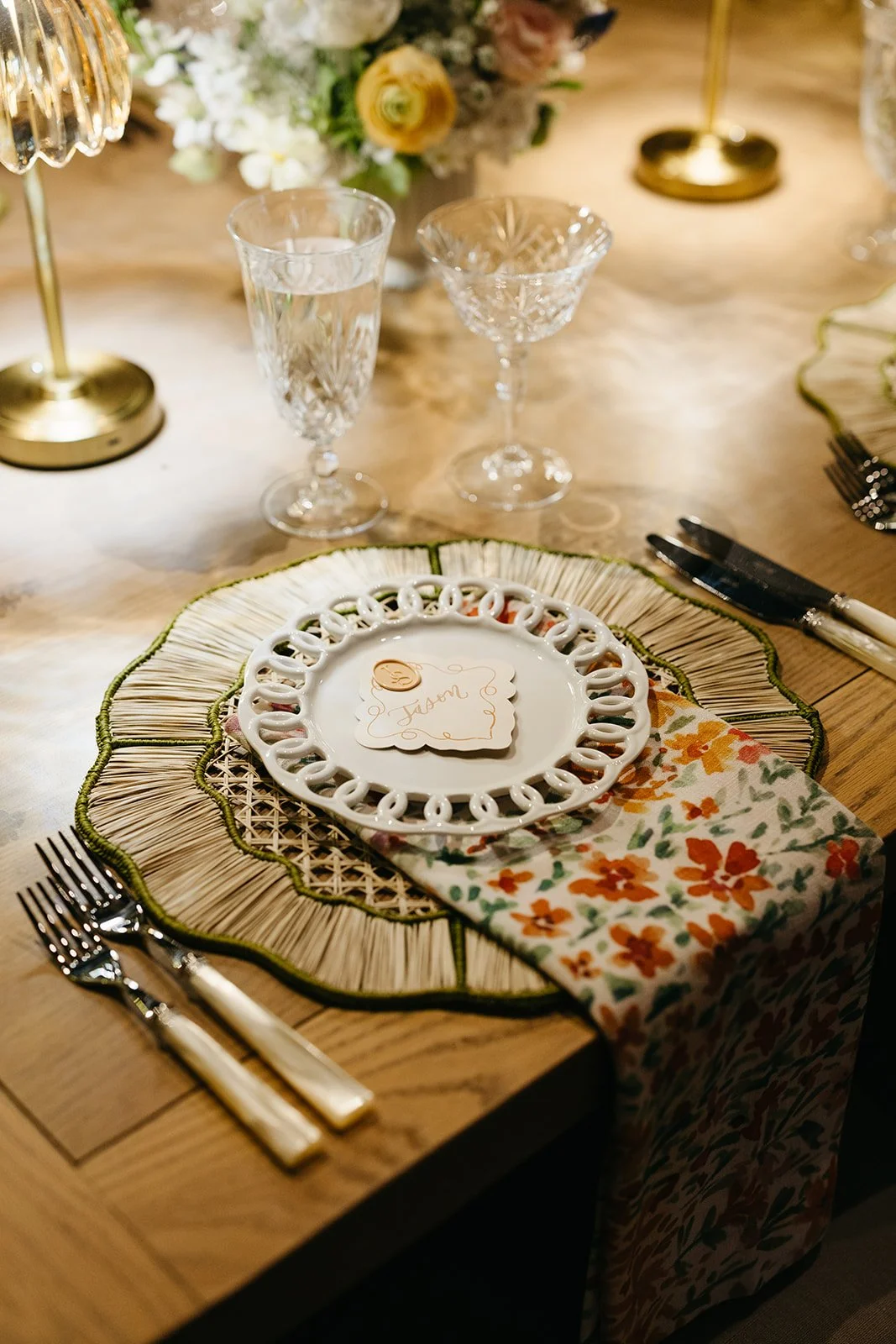 A table setting with a decorative charger plate, a floral napkin, a place card, wine glass, water glass, cutlery, and a floral centerpiece in the background.