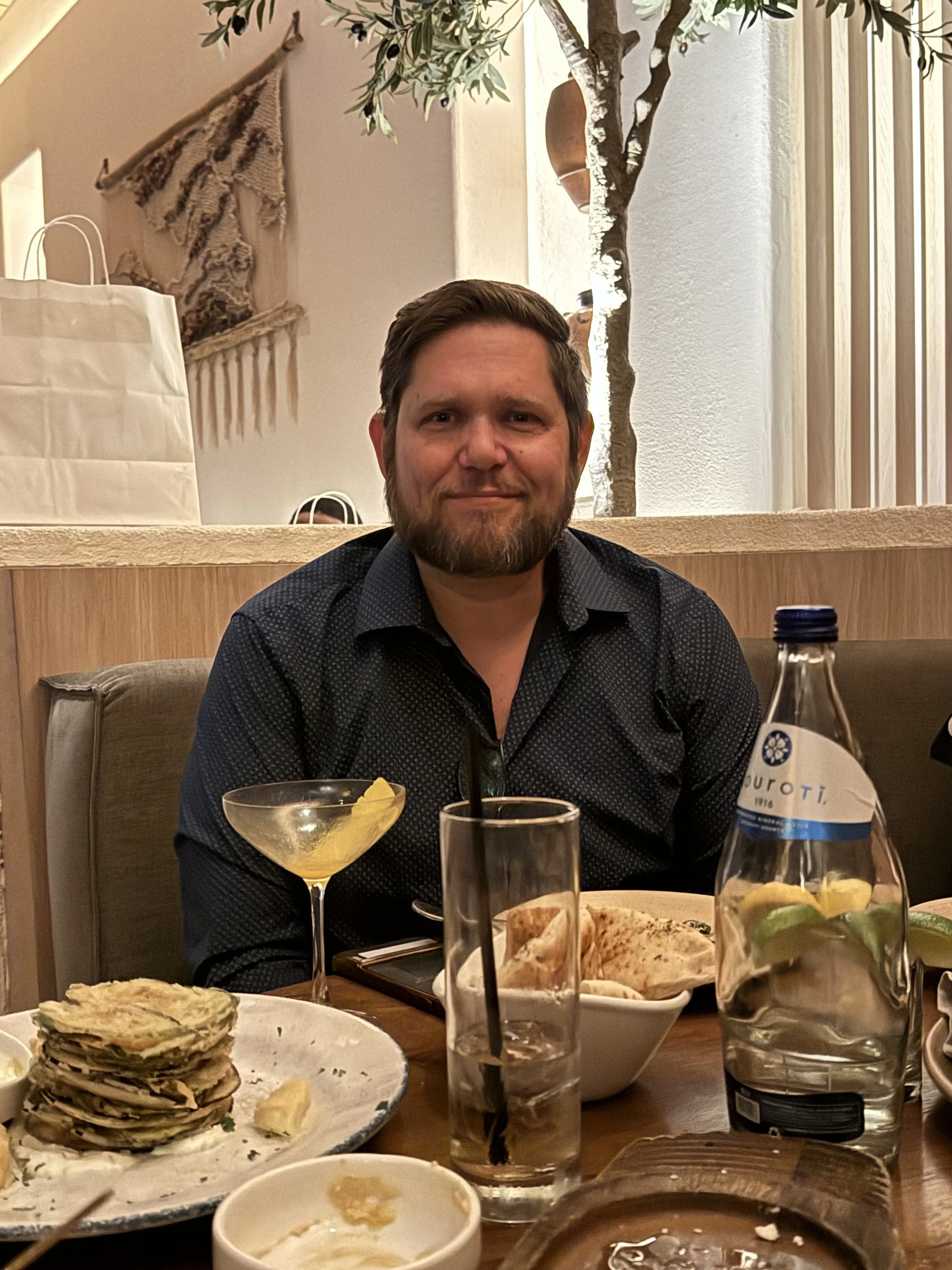 A man sitting at a restaurant table with food and drinks, smiling at the camera. The table has a stack of quesadillas, a cocktail, a water bottle with lime, and other dishes. The background features a light-colored wall, a plant, and decorative hangi
