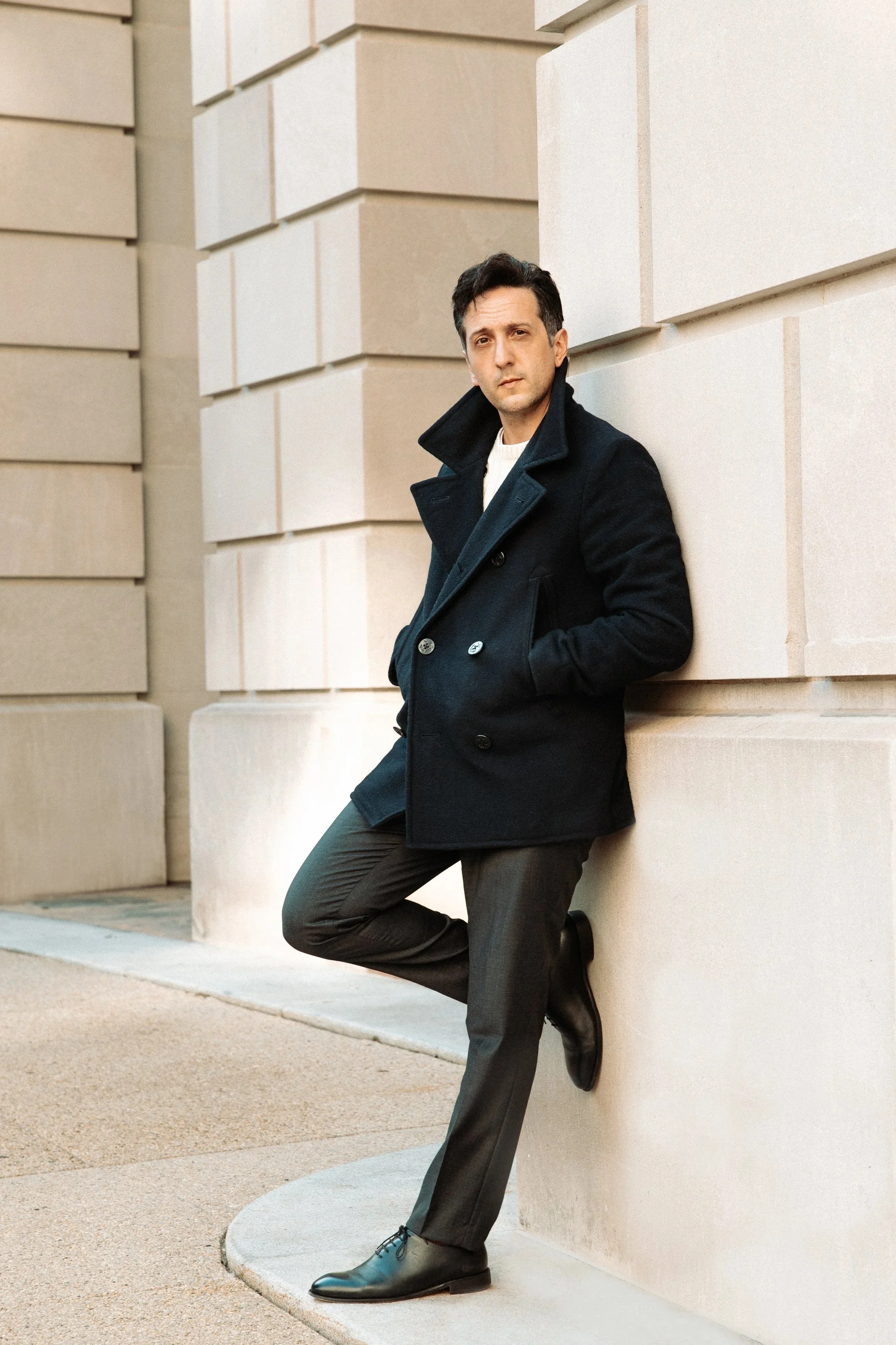 A man in a black suit holding a glass of red wine, standing in a dimly lit modern setting.