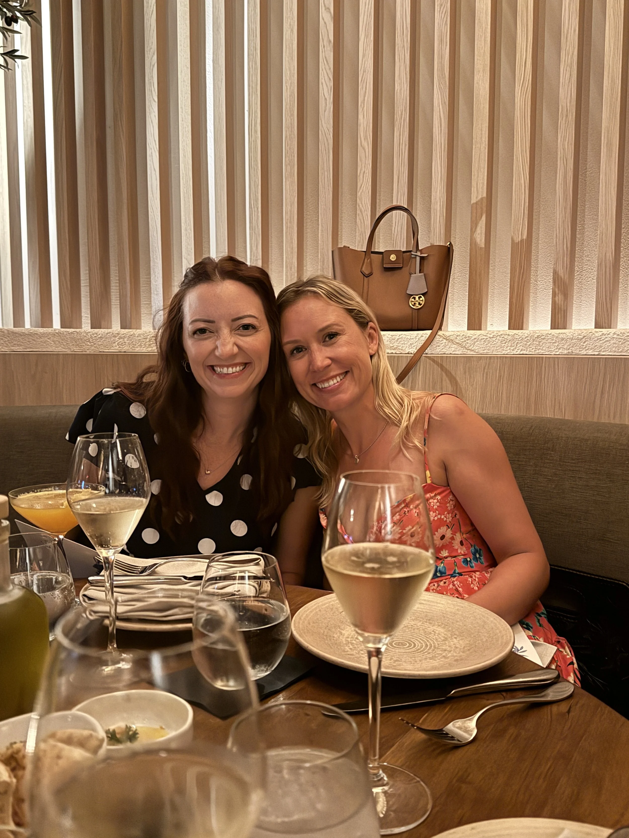 Two women sitting at a restaurant table, smiling, with drinks and dinnerware in front of them, and a beige handbag hanging on the wall behind them.