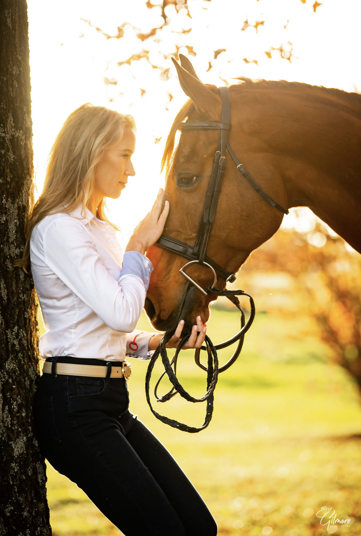 Woman smiling next to a brown horse with a background of trees and sunlight.
