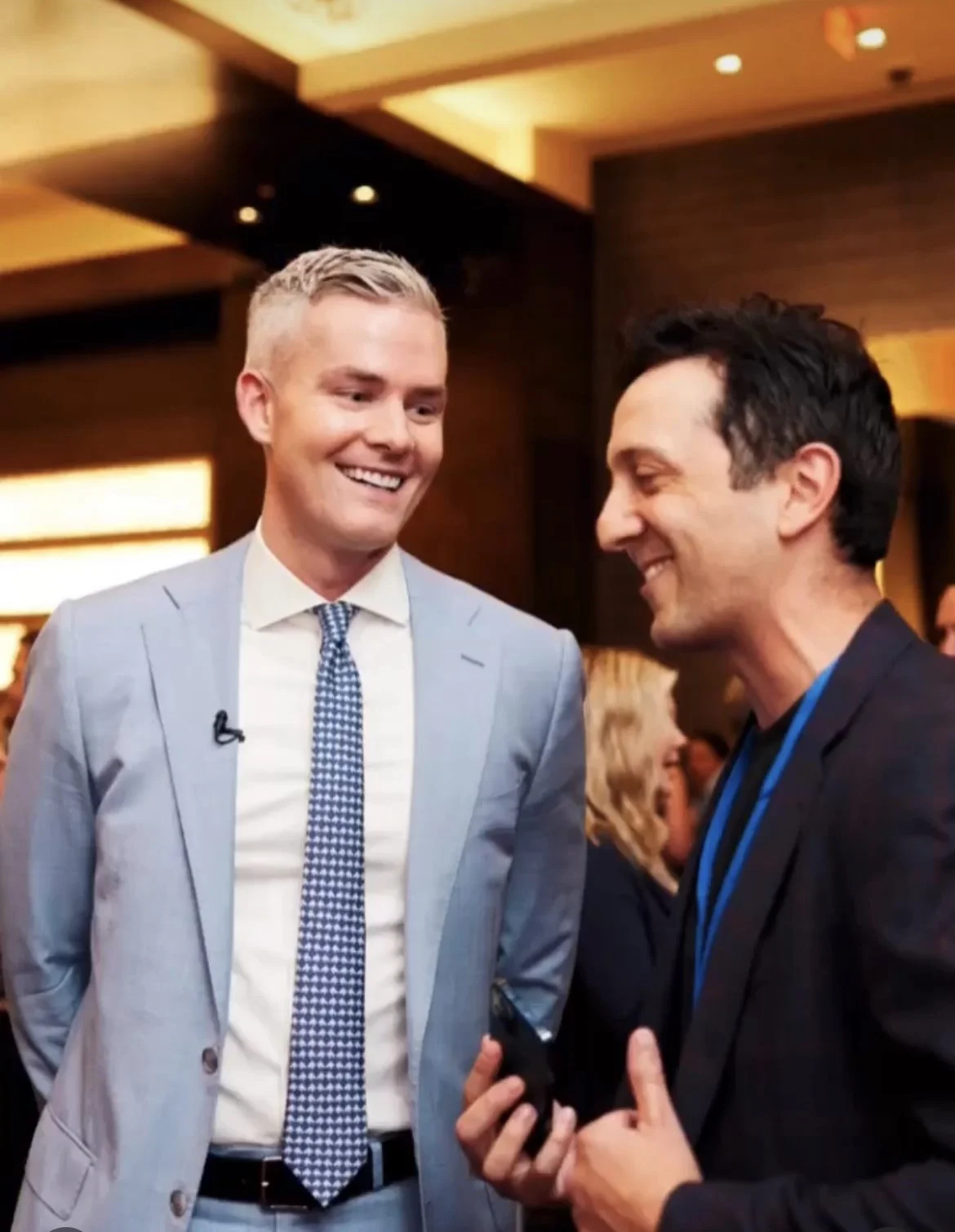 Two men dressed in formal business attire smiling and talking at an event, with a woman blurred in the background.