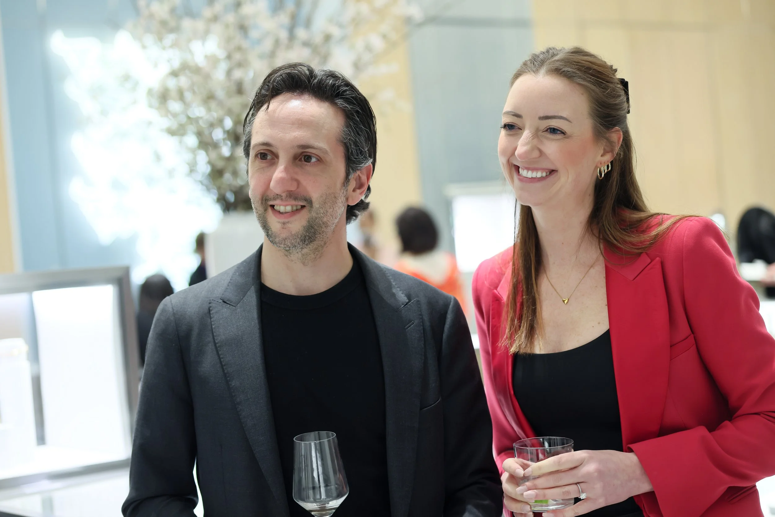 A man and woman smiling at a social event, holding drinks, with blurred people in the background.