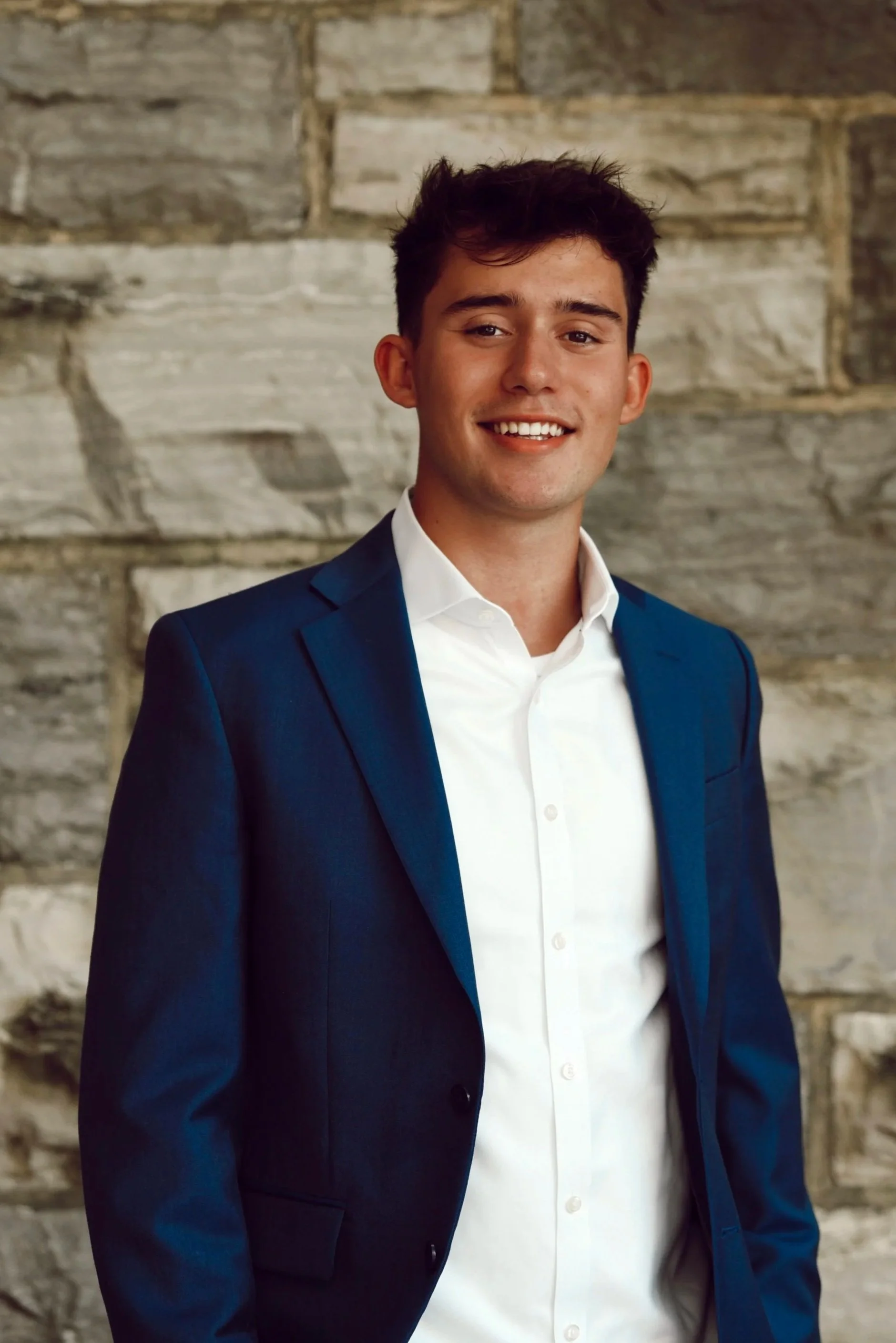 Young man with dark hair and a smile, wearing a green jacket over a beige sweater and gray shirt standing outdoors in front of a brick building during what appears to be late afternoon or early evening.