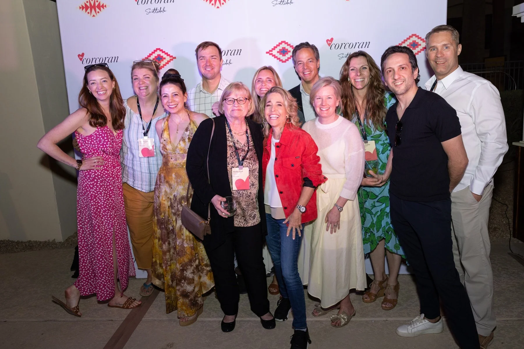 Group of people at a social event, standing in front of a backdrop with the logos 'Be a Corcoran' and 'Corcoran Scottsdale', smiling and posing for a photo.