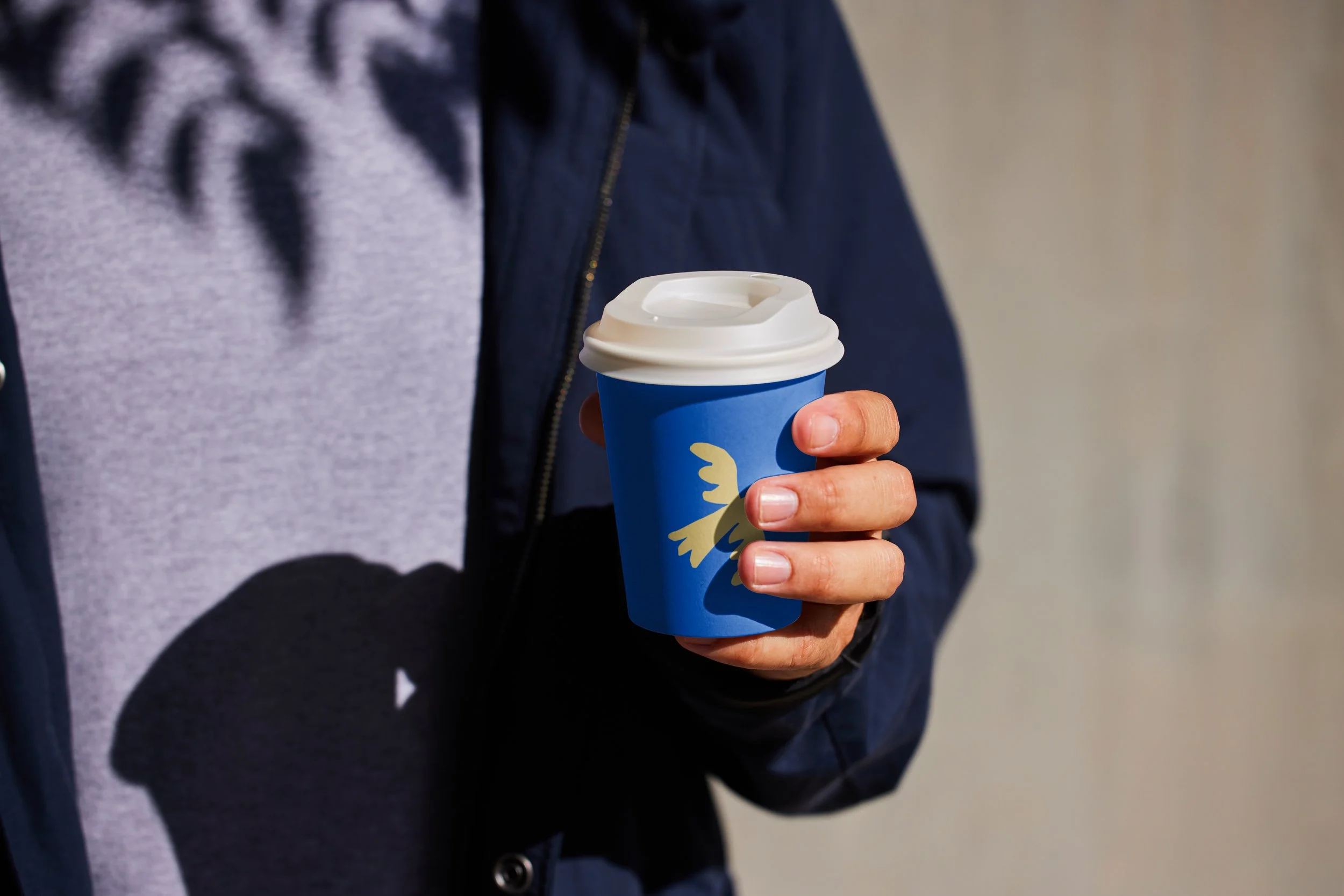 Une personne tient une tasse à café bleue avec un logo d'oiseau beige, portant un couvercle blanc, en plein jour.