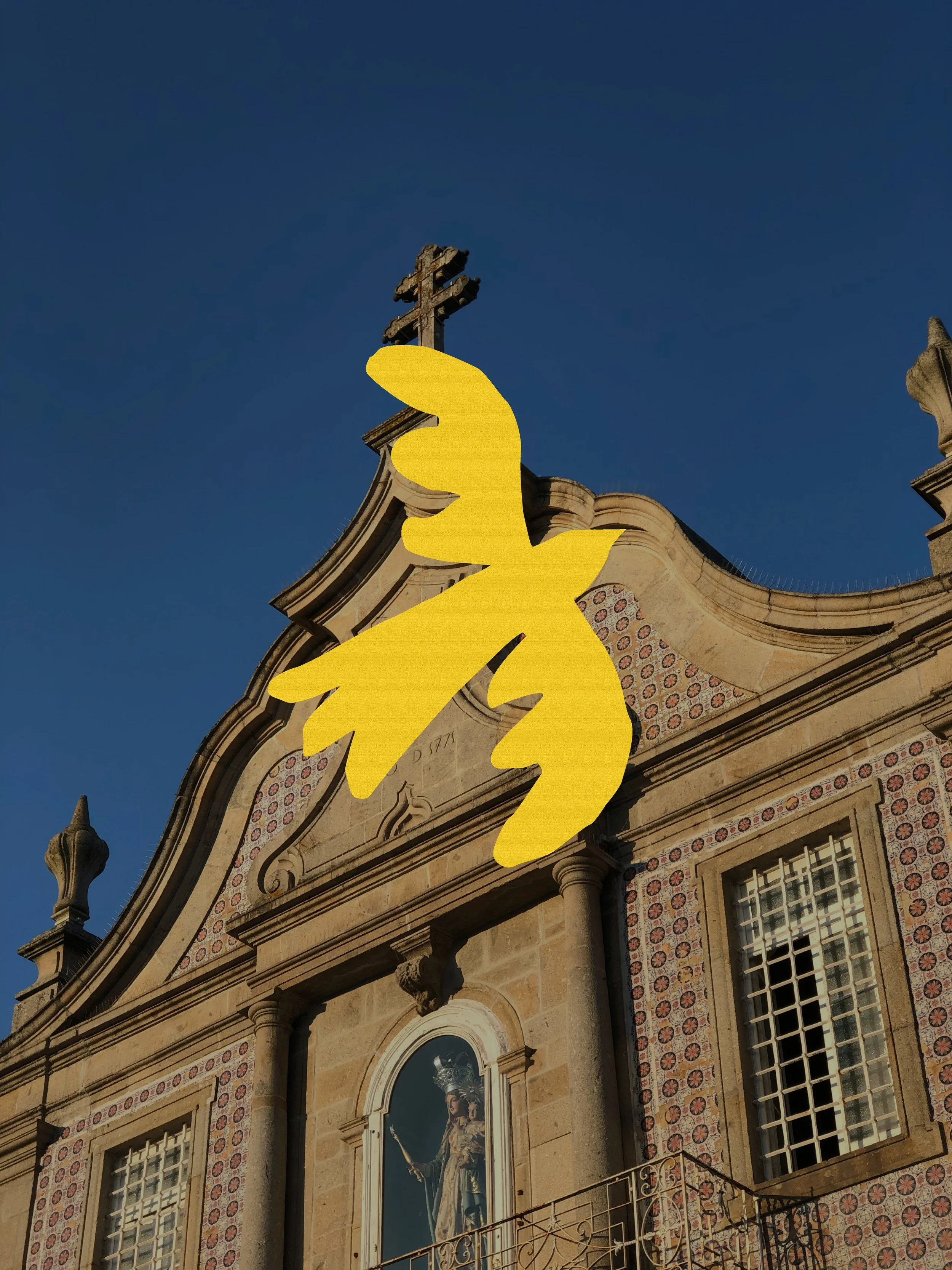 Façade d'une ancienne église ou cathédrale en pierre avec une statue religieuse dans une niche, des fenêtres à carreaux et une croix en pierre au sommet, en plein jour avec un ciel bleu clair.