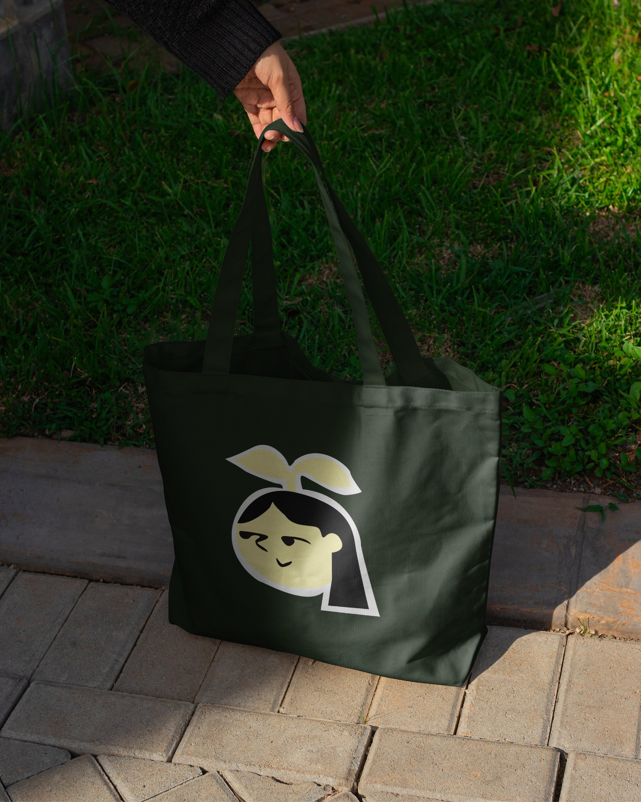 Un sac vert avec une illustration d'une fille souriante avec des cheveux longs et une feuille sur la tête, tenu par une personne dehors sur un trottoir en pavés, près d'un jardin.
