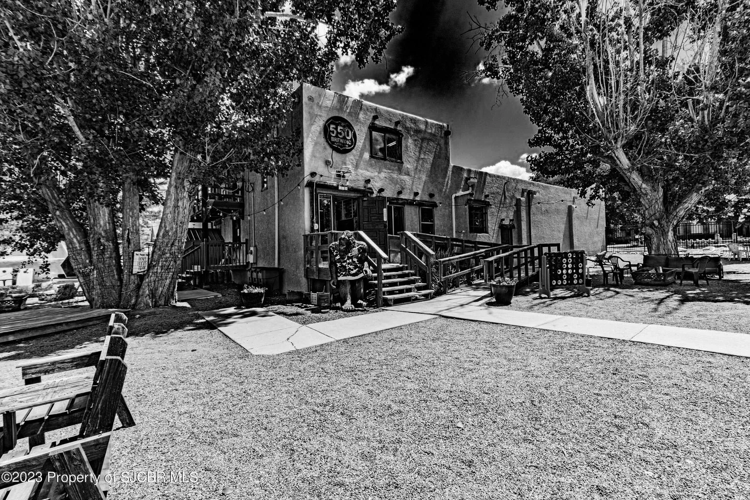 Black and white photo of a two-story building with a sign that reads '550 Brewing'. The building has a small set of stairs at the front and an outdoor seating area with benches and tables. There are large trees with thick trunks and many leaves around the building, providing shade.