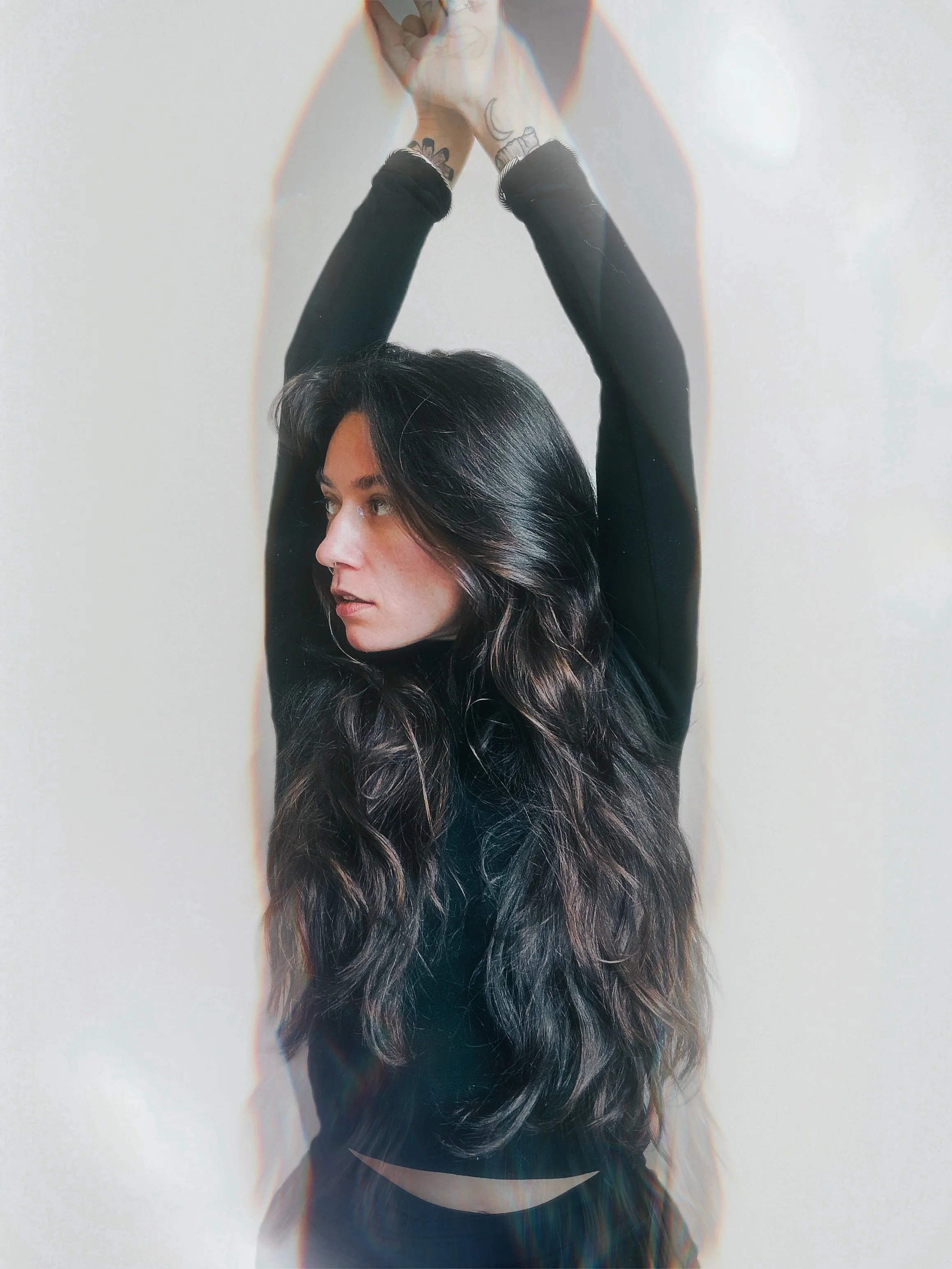 A woman with long, wavy dark hair extending downward, wearing a black long-sleeve top, extending her arms upward with her hands clasped together, standing against a plain wall.