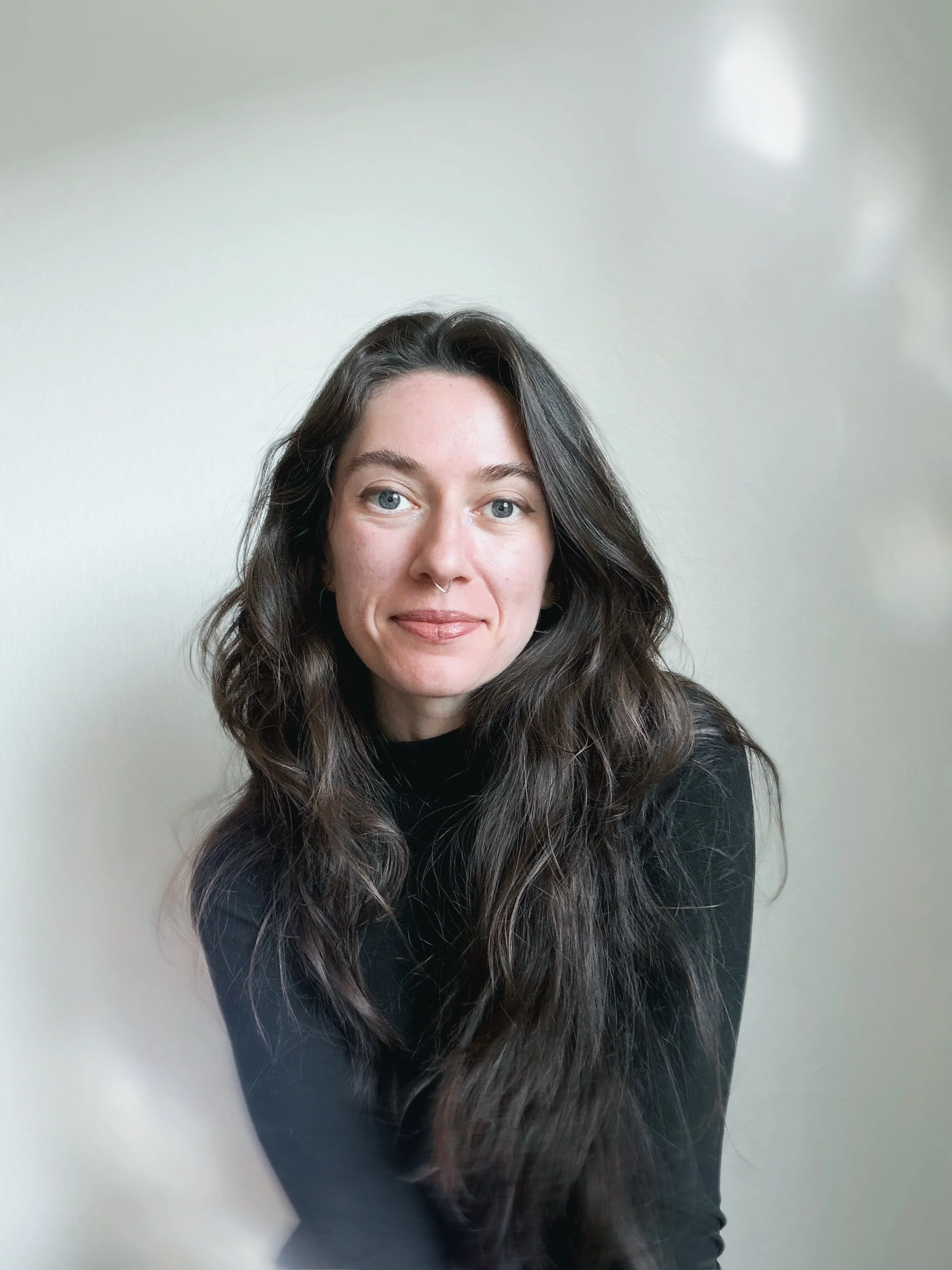 A woman with long wavy dark hair, light skin, and blue eyes, wearing a black top, smiling gently against a plain light-colored wall.