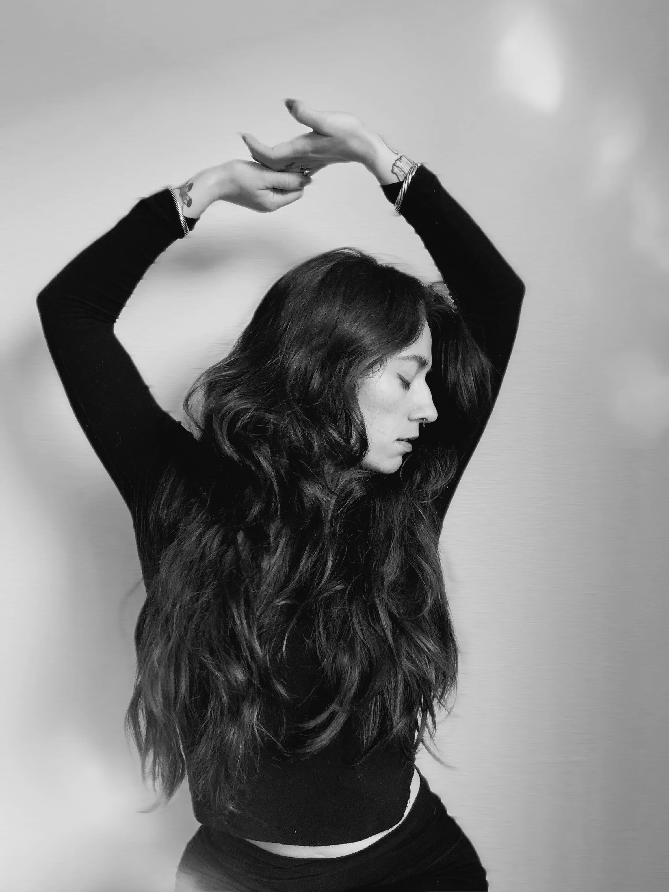 A woman with long wavy hair standing against a light-colored wall, with her eyes closed and arms raised above her head in a relaxed pose.