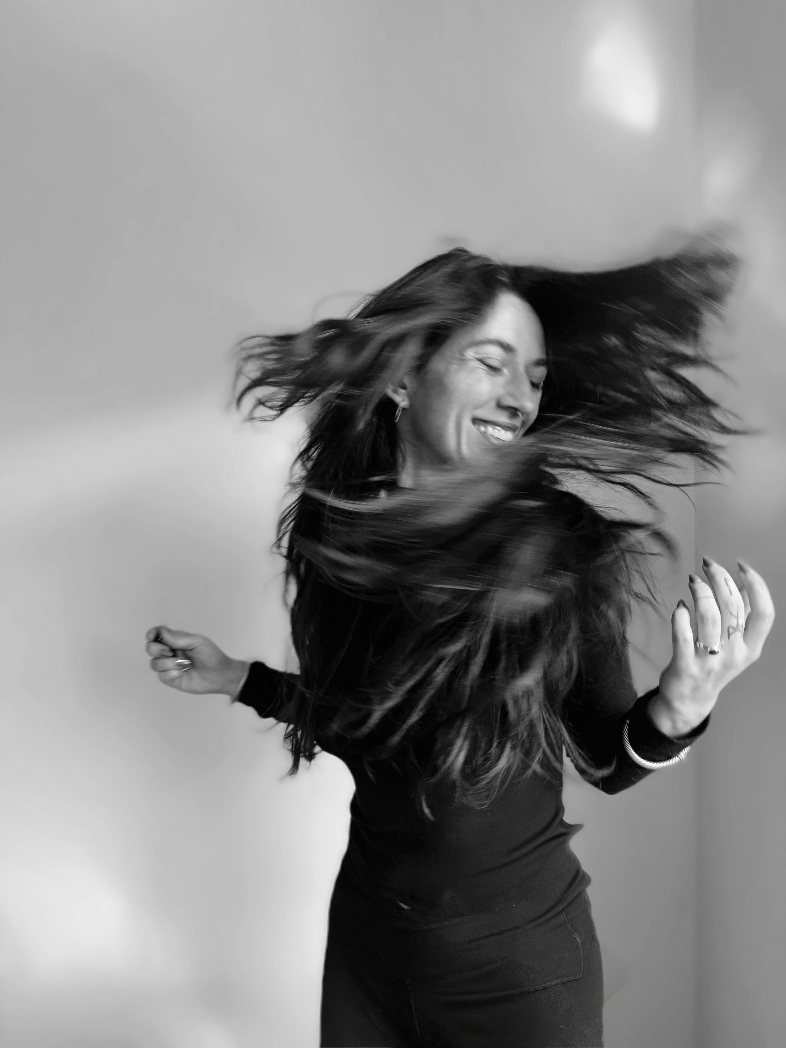 Black and white photo of a woman with long hair, smiling with eyes closed, hair flying. She is wearing earrings and a long-sleeve top, holding her right hand with rings and a bracelet, against a plain background.