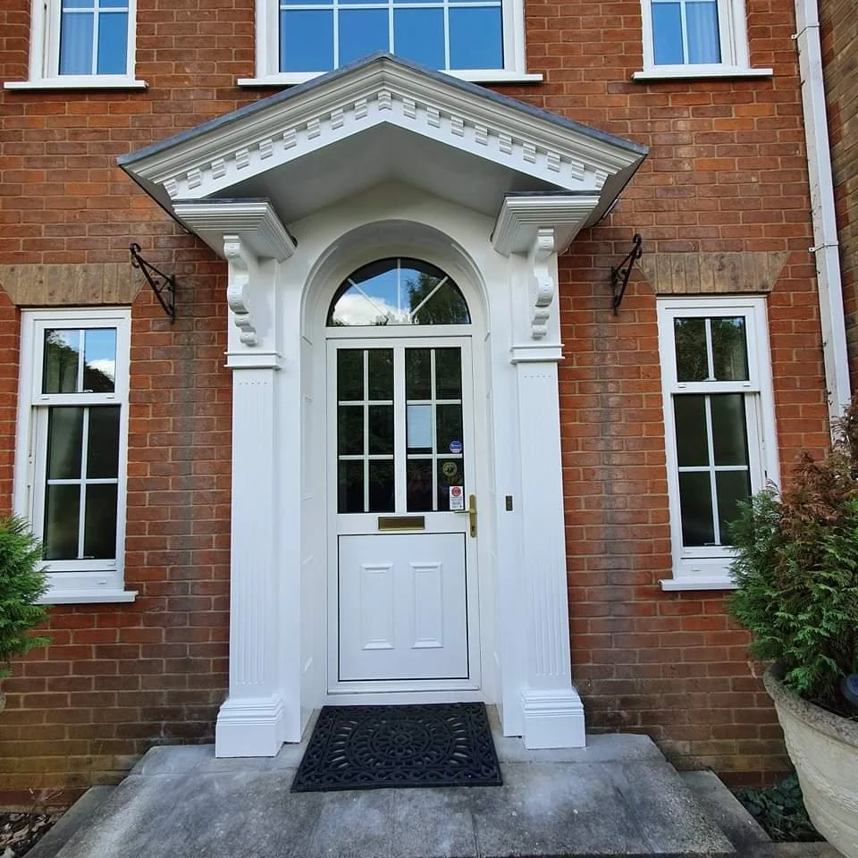 Traditional brick house entrance with white front door and decorative portico, flanked by windows with white frames.