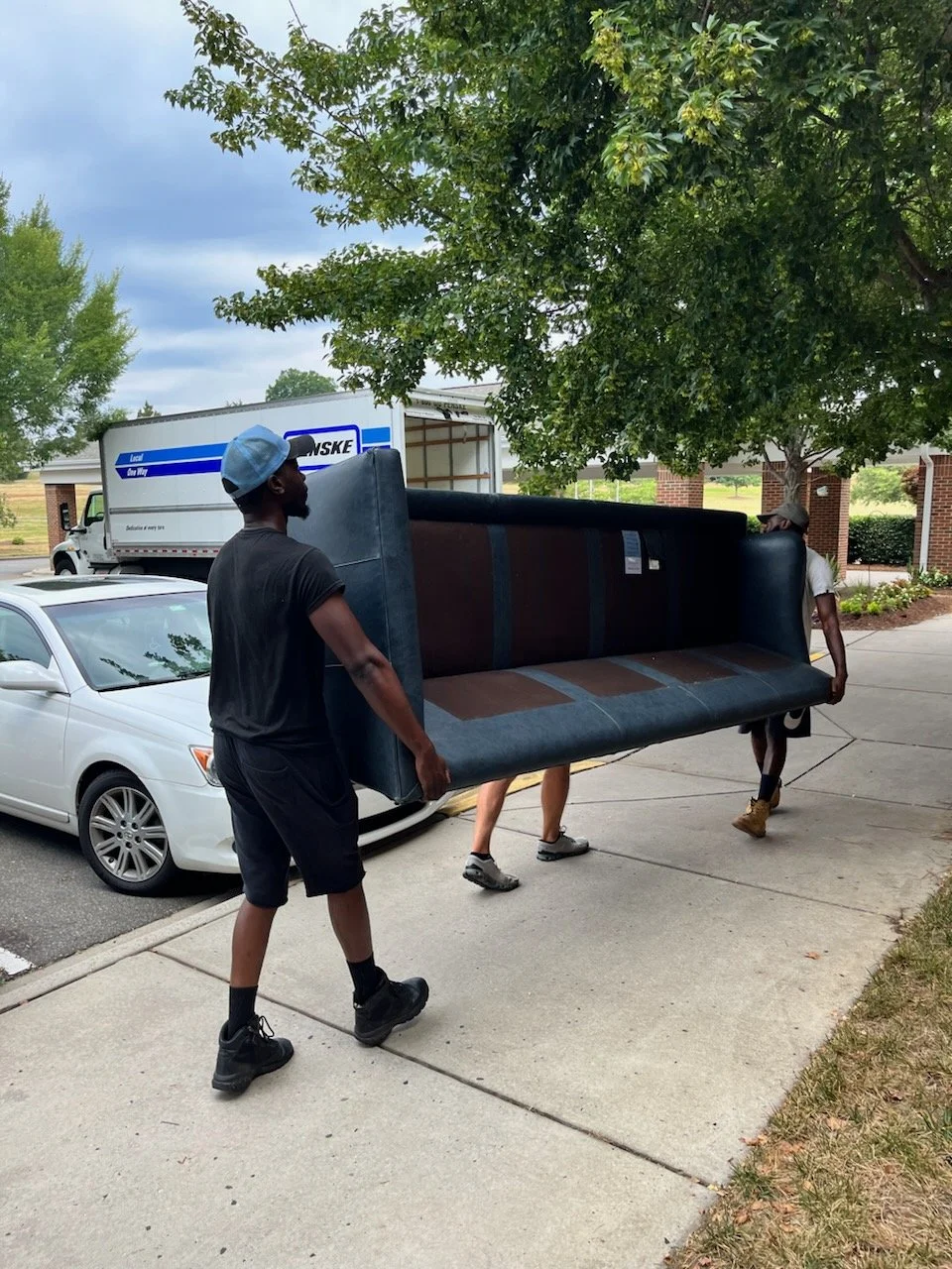 Three men who work for th moving company Epoch Movers are moving a dark-colored couch across a sidewalk near parked cars and a tree in a suburban area.