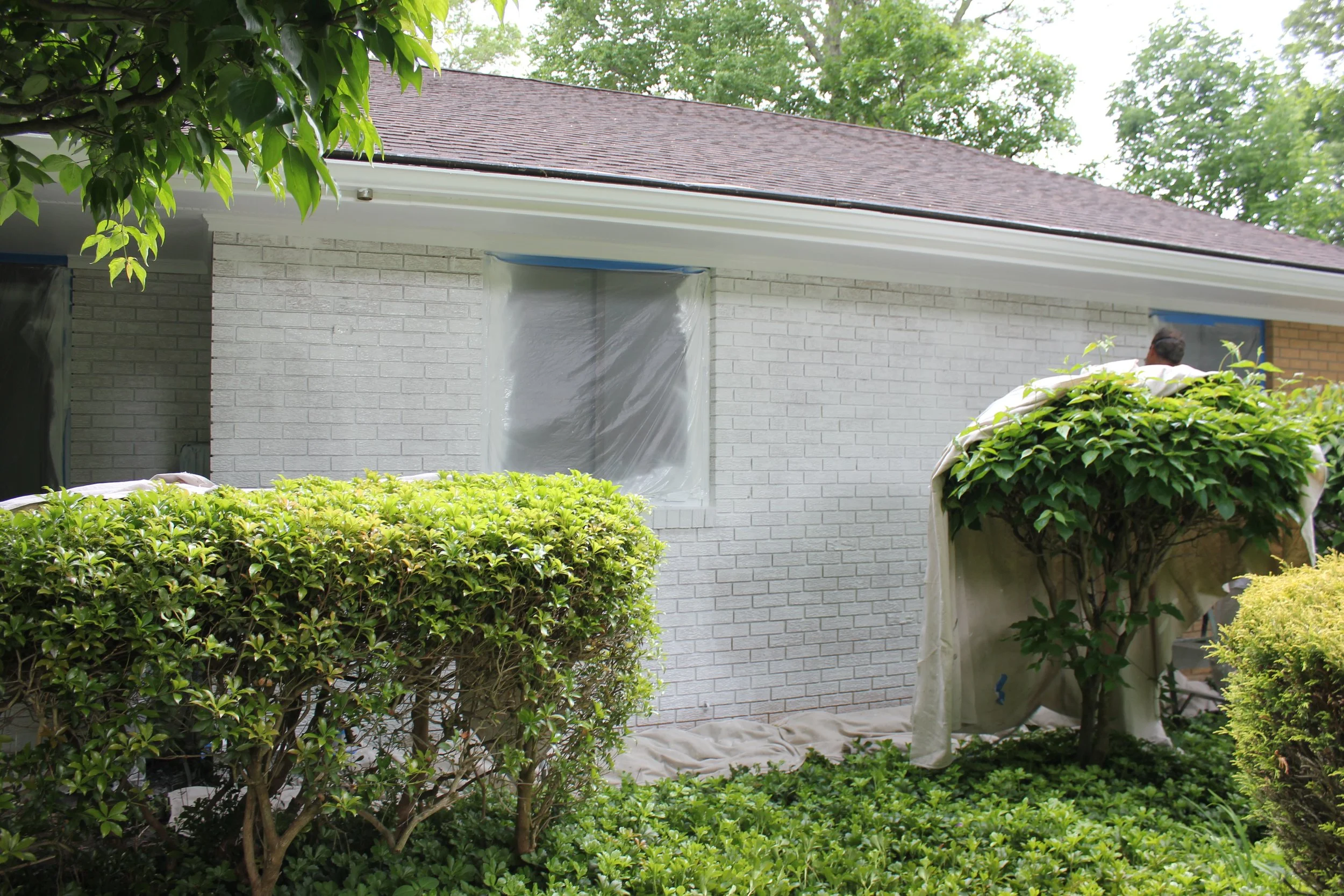 The exterior of a house with white brick walls, a brown shingle roof, and a lawn with bushes. There is some fog or dust covering part of the wall and window, and a person is partially visible behind a bush.
