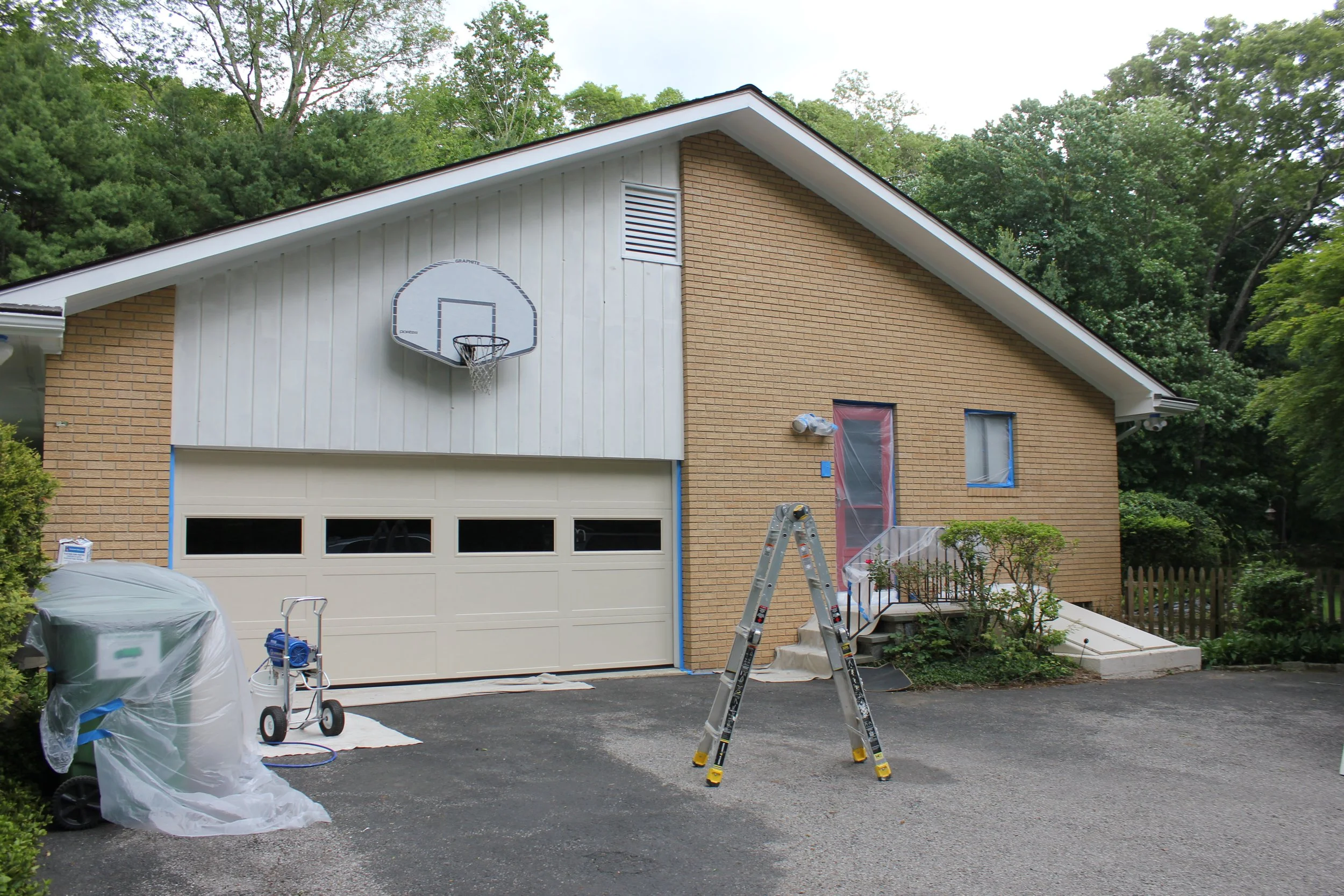 Home exterior undergoing renovation, with a basketball hoop on the garage and construction tools like a step ladder, plastic sheeting covering work areas, and a ladder in the driveway, surrounded by green trees.
