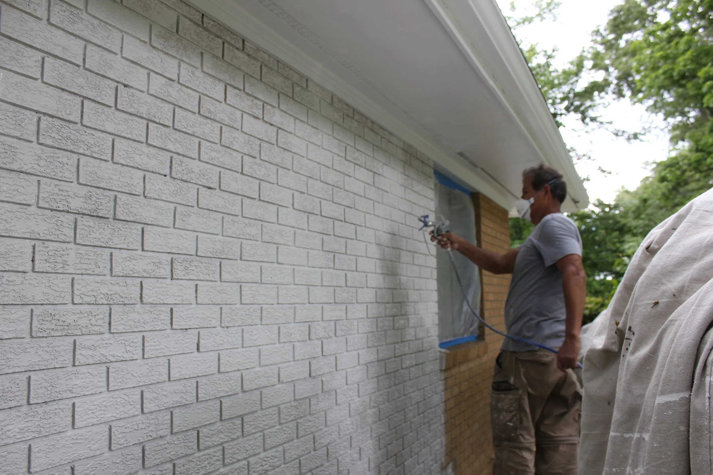 A man spray painting a white brick wall outdoors. He is wearing a gray t-shirt and a face mask, with trees visible in the background.
