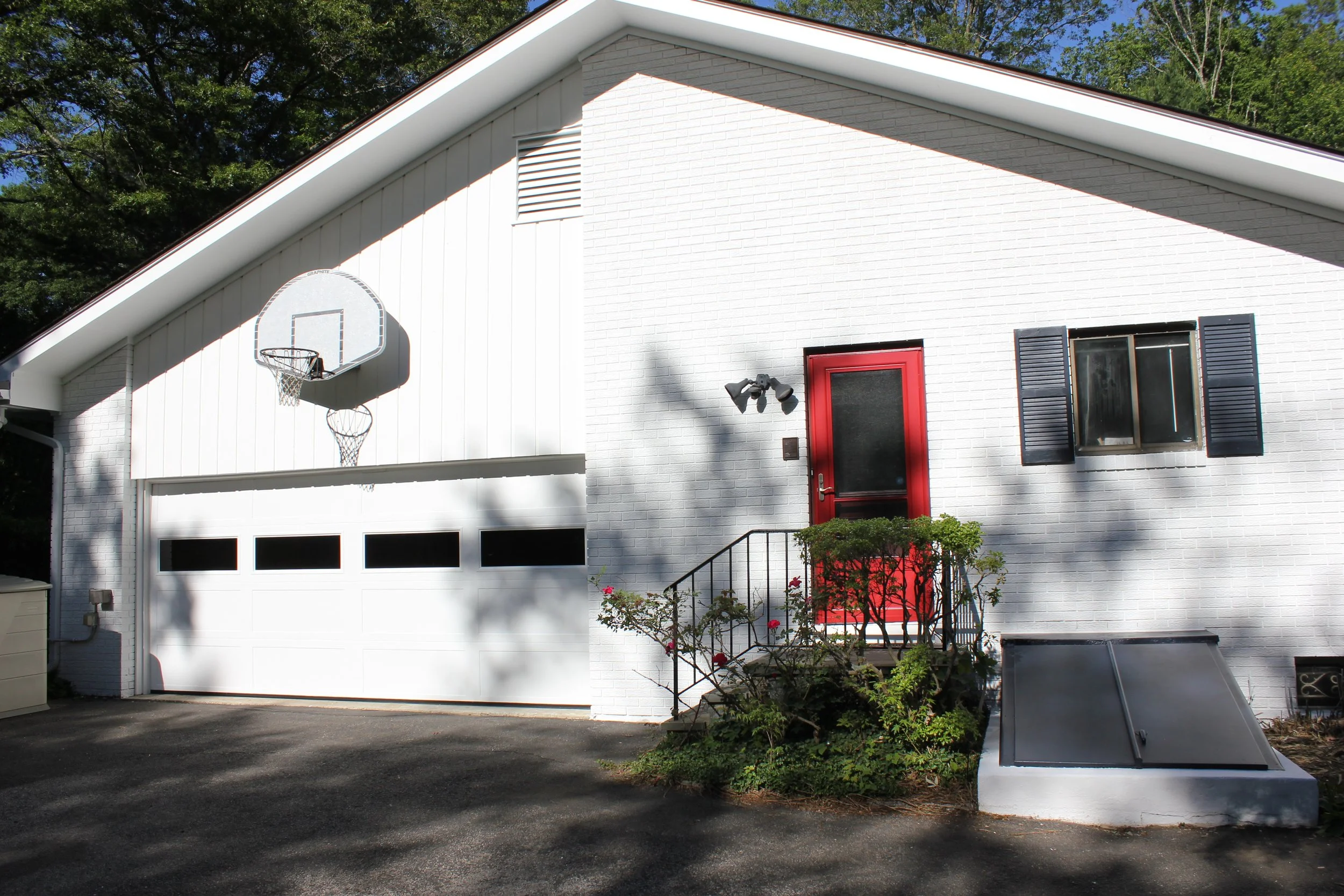 White house with a red door and black shutters, a basketball hoop above the garage, solar panels on the ground, and greenery in front, with a background of tall trees.