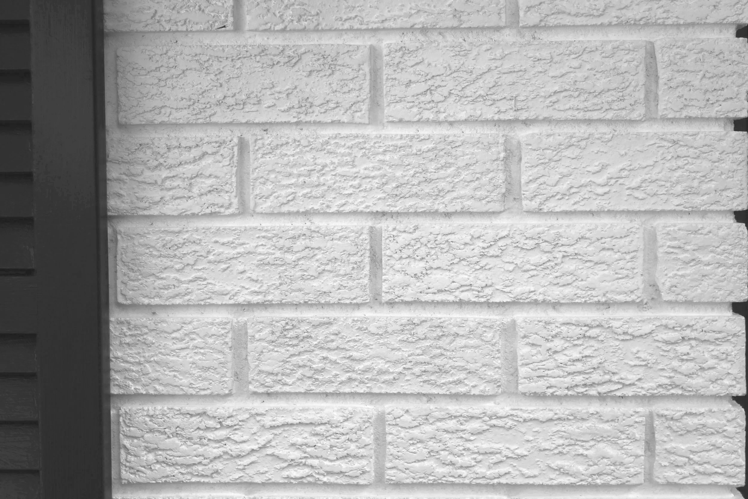 Close-up of a white brick wall with textured surface, showing horizontal mortar lines, and part of a dark-colored door frame on the left side.