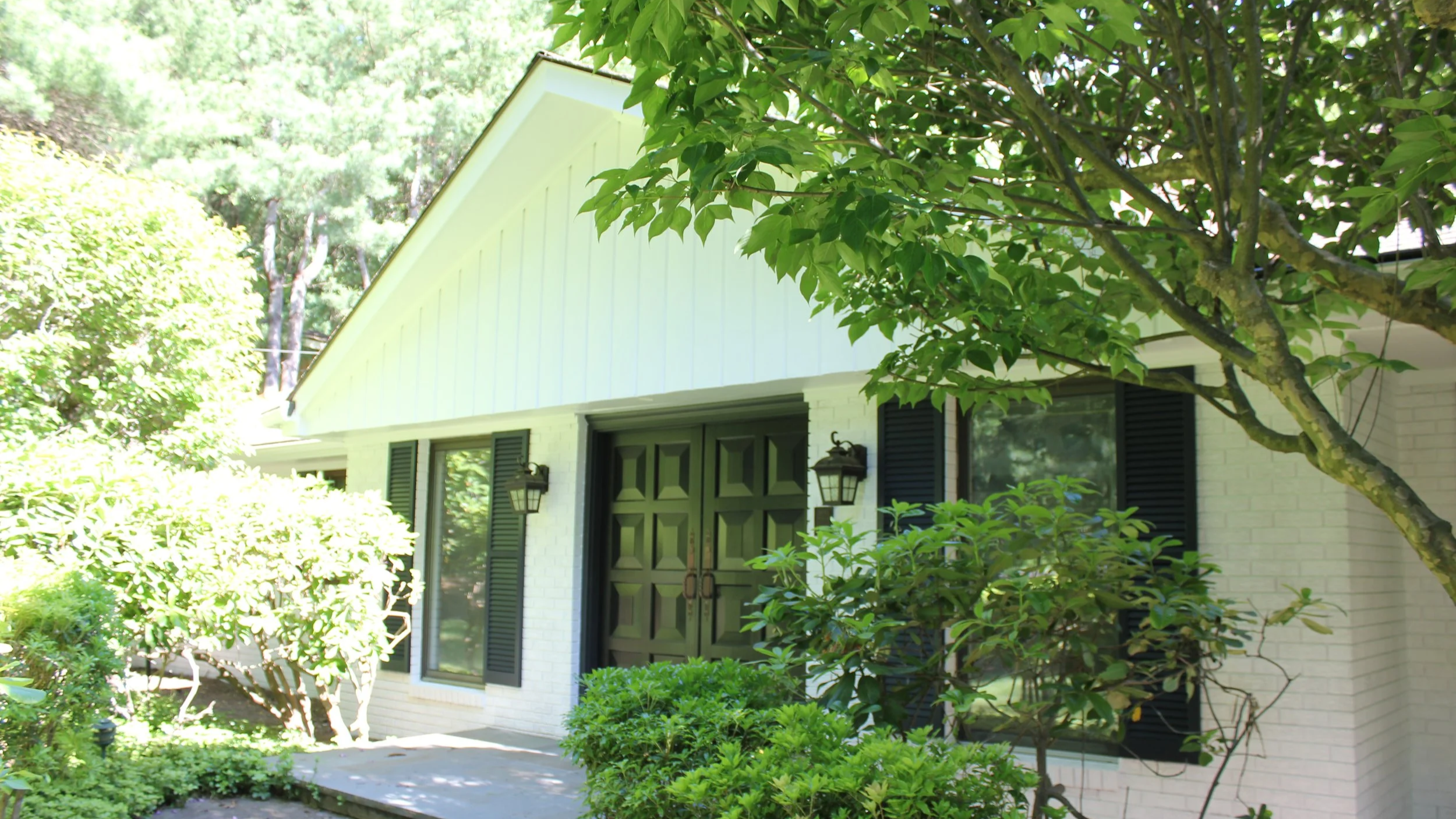 Front view of a house with a white brick exterior, green double doors, black shutters, and black outdoor lanterns, surrounded by green bushes and trees with a wooded area in the background.