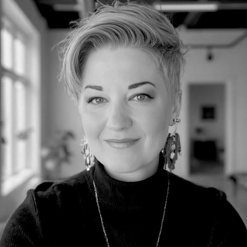 A woman with short, styled hair and earrings, smiling indoors with natural light coming from windows.
