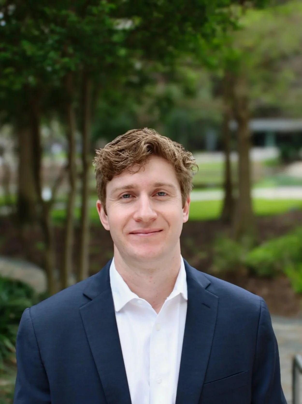A young man in a navy blue suit and white shirt stands in a room with exposed brick wall, wood door, and plain beige wall behind him.