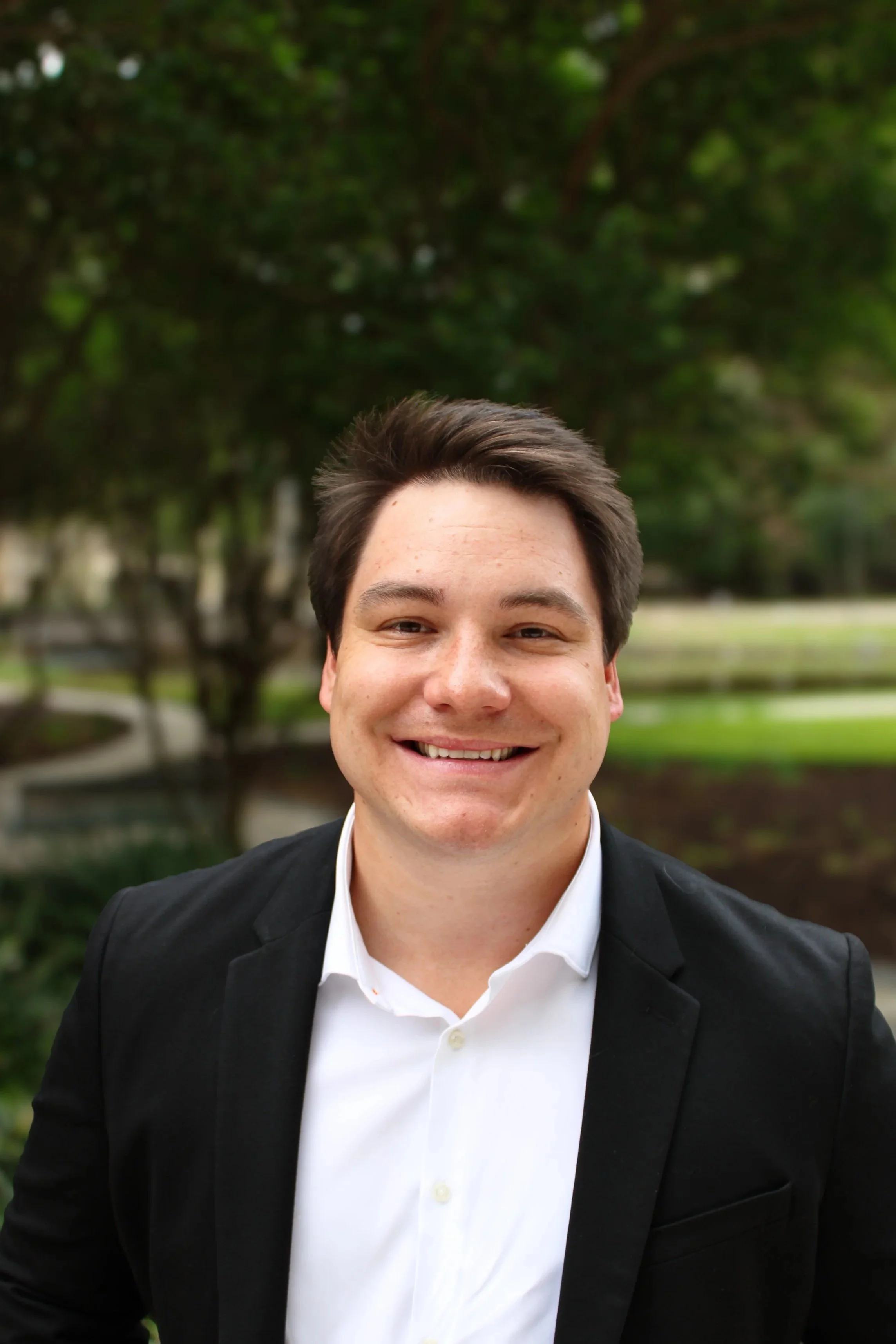 A young man in a business suit standing indoors near colorful framed posters, with a potted tree in the background.