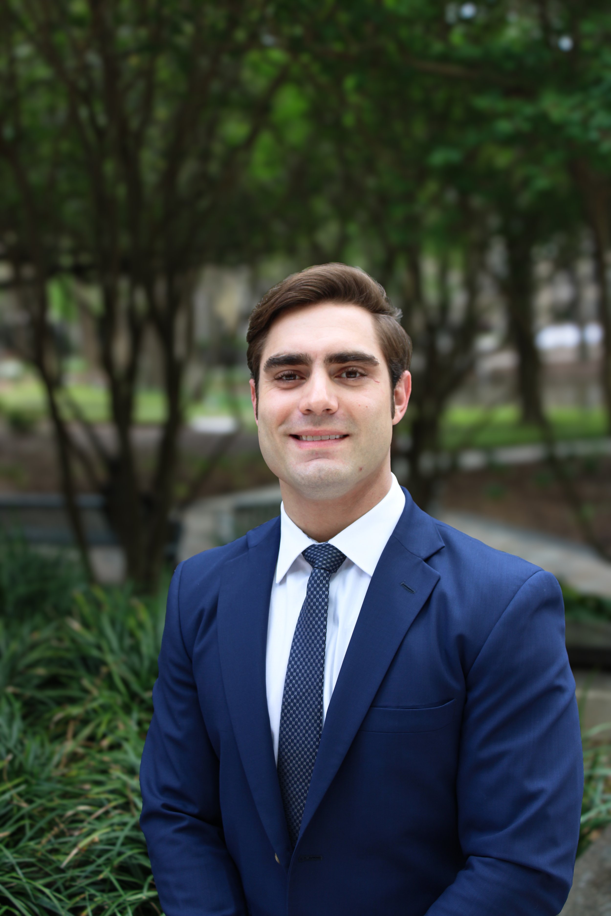 A man in a blue checkered blazer and white shirt standing outdoors next to a brick wall, with a sidewalk, parked cars, and potted plants visible in the background.